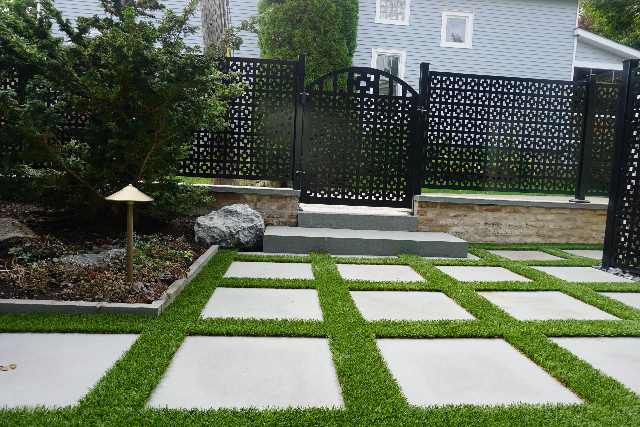 A backyard with a black decorative metal fence, a small mailbox, paver stones on the grass, a large tree, shrubs, rocks, and a house with light blue siding.