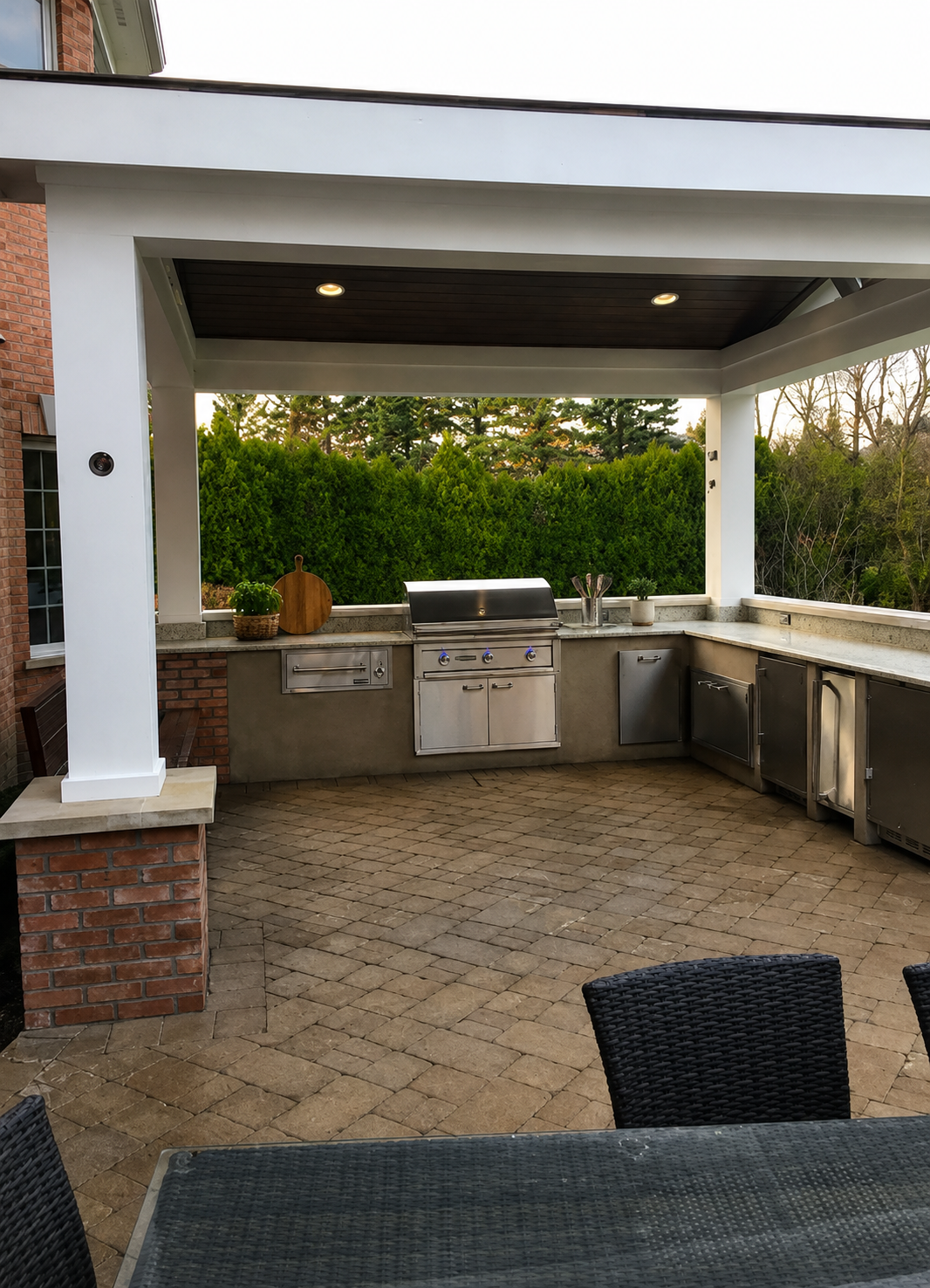 Outdoor kitchen with stainless steel grill, countertop, and storage cabinets under a covered patio, surrounded by green trees and brick features.