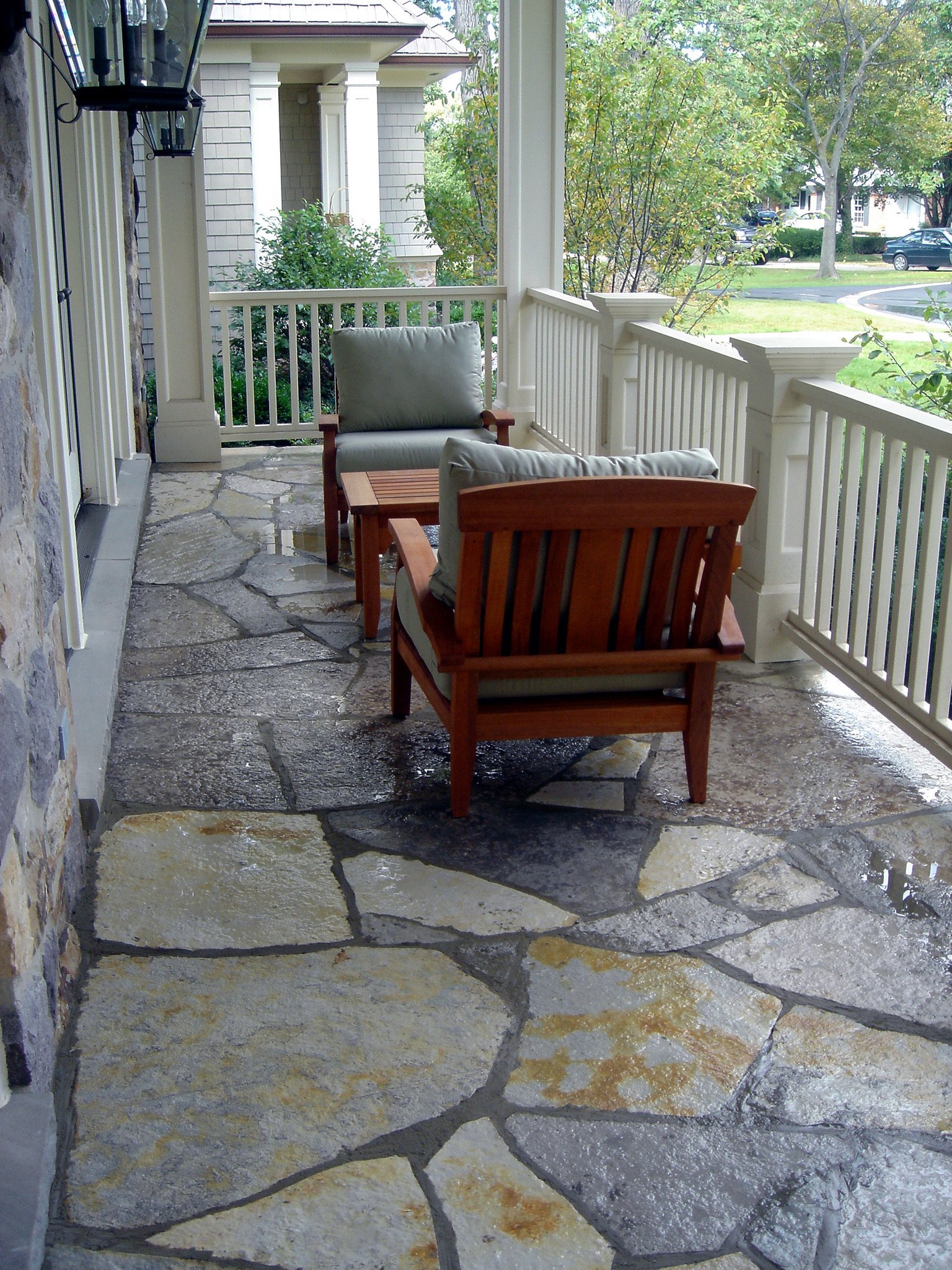 A porch with a stone floor, featuring two cushioned wooden chairs and a small wooden table, surrounded by a white railing and greenery outside.