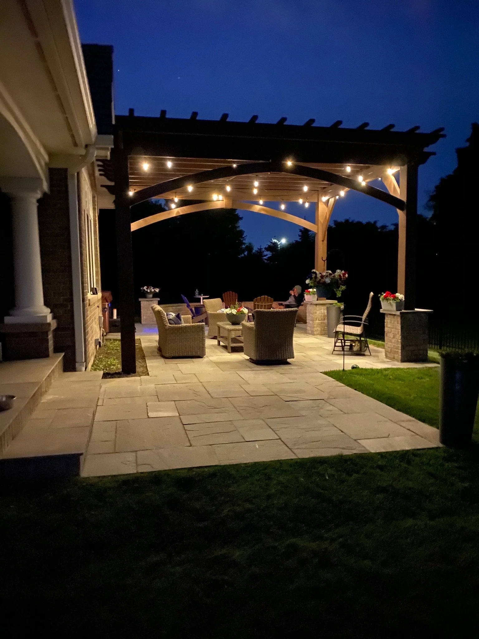 An outdoor patio at night with string lights hanging under a wooden pergola, surrounded by flowers and chairs, with a person sitting and a dark sky in the background.