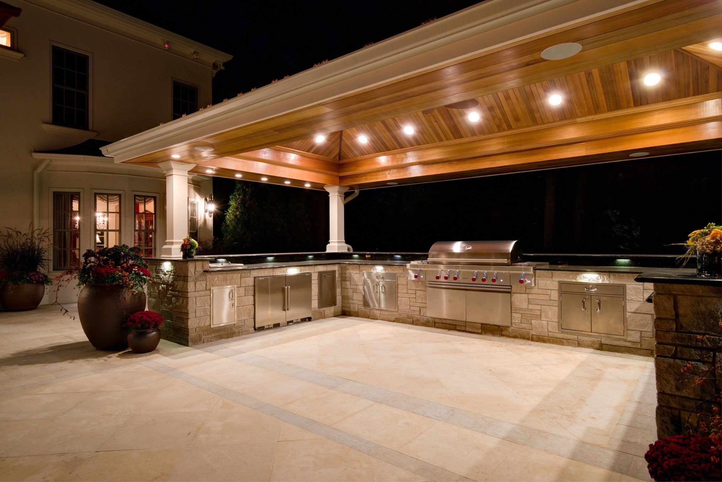Night view of an outdoor kitchen area with a brick island, stainless steel grills, a wooden roof structure with embedded lights, potted plants, and an adjacent house with lit windows.