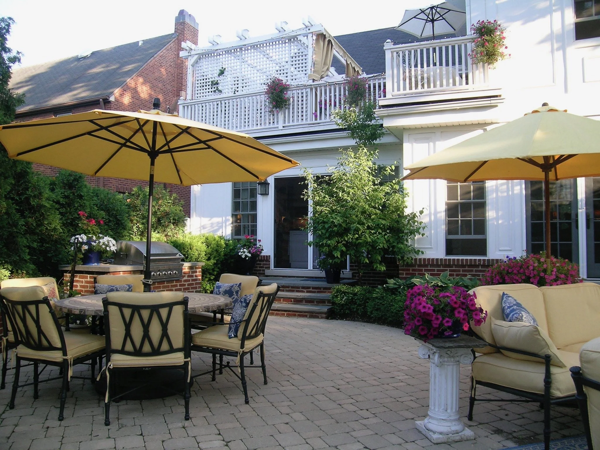 A backyard patio with yellow umbrellas, garden furniture, potted flowers, and a white house with a balcony and large windows.