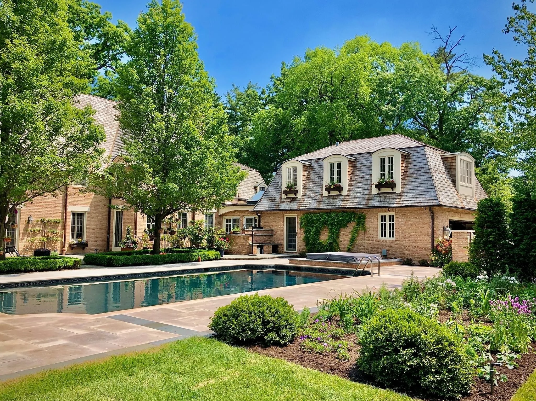 A luxurious backyard with a swimming pool, surrounded by lush green trees and well-maintained gardens, featuring a two-story brick house with dormer windows and a shingled roof.