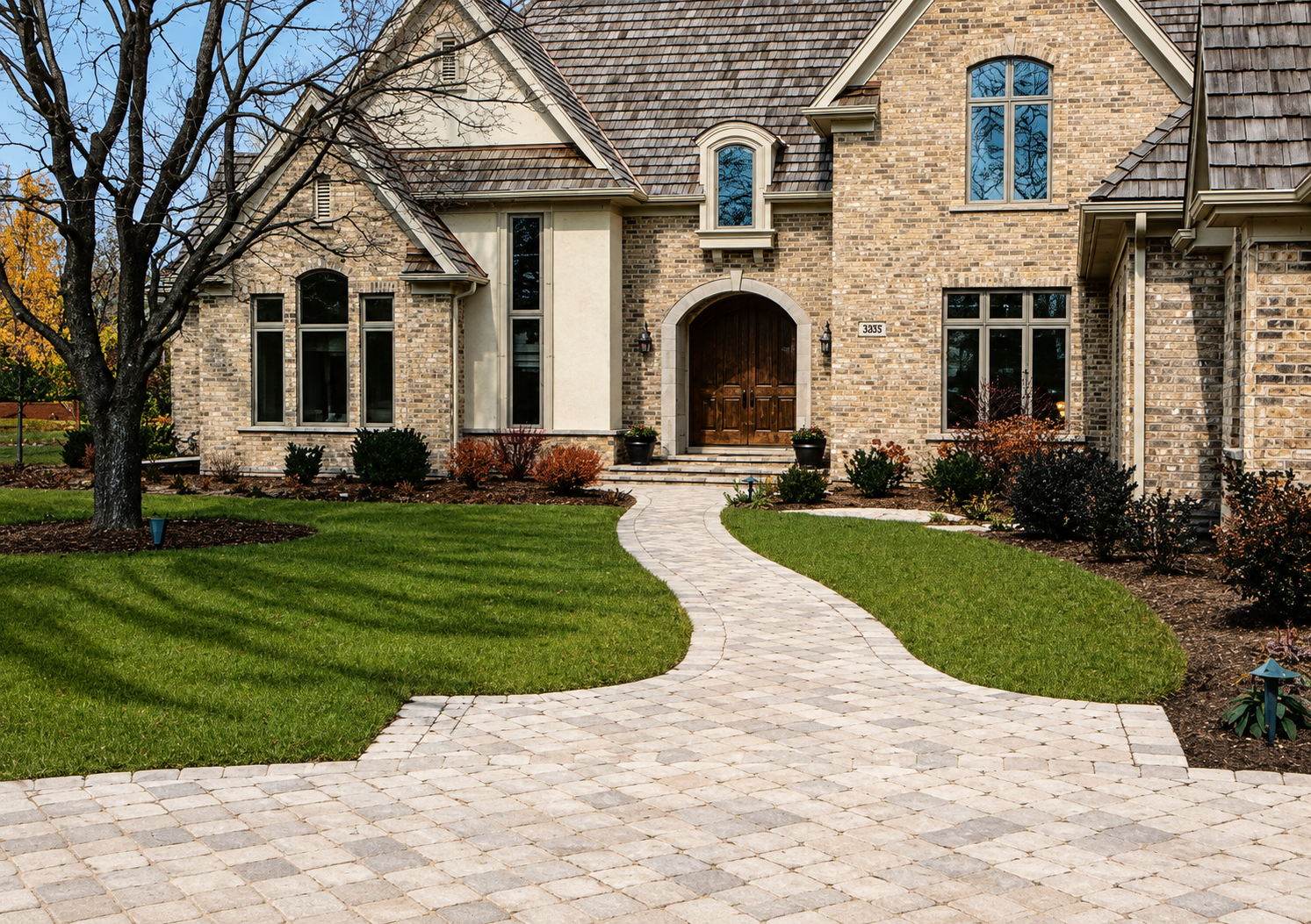 Front view of a large brick house with a curved stone walkway leading to a wooden front door, surrounded by a well-kept lawn and landscaped garden with bushes and trees.