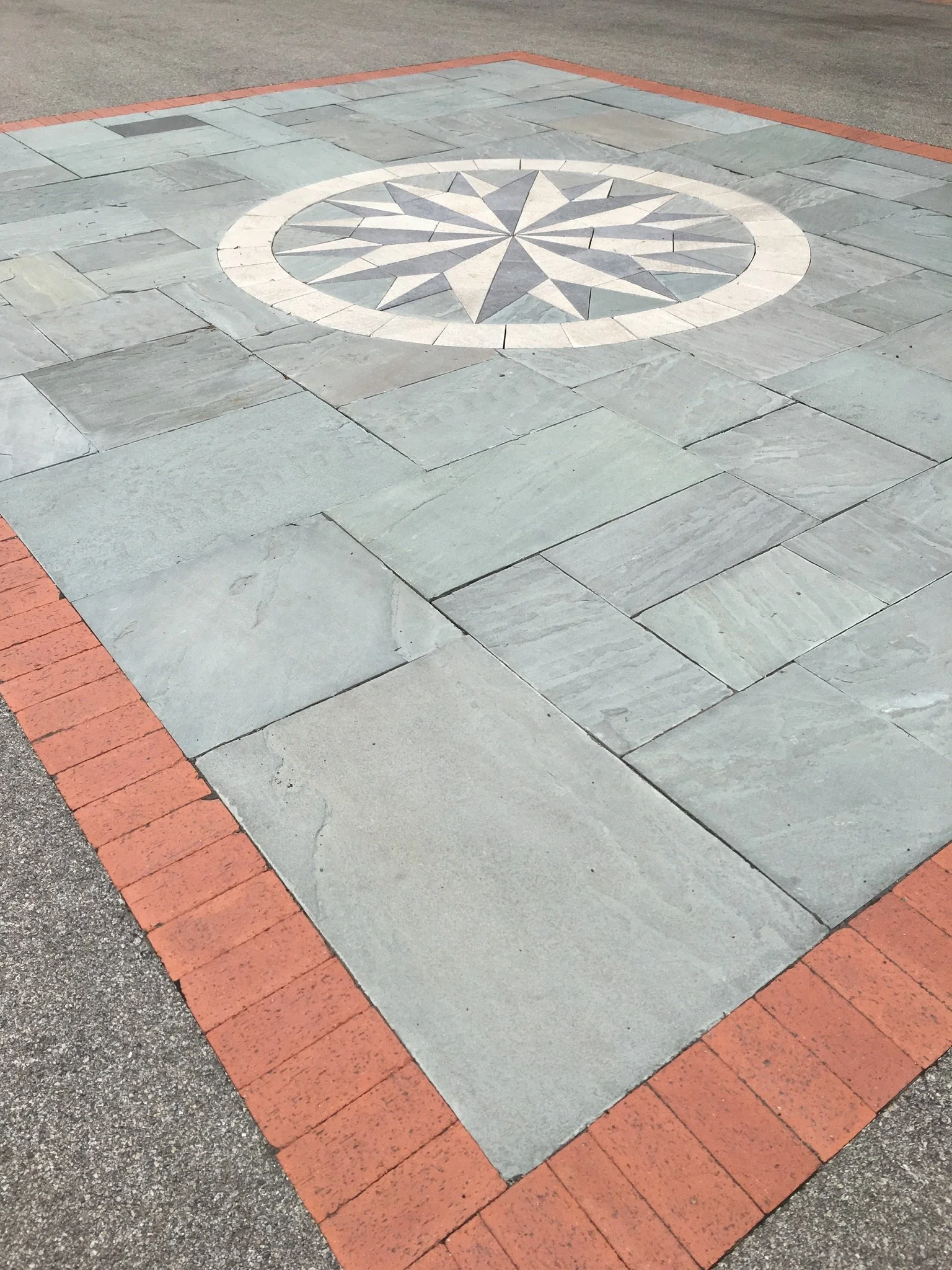 A decorative circular pattern on a paved area, featuring a starburst design with a mix of light and dark gray stones, surrounded by a white border, and bordered by a red brick edge.