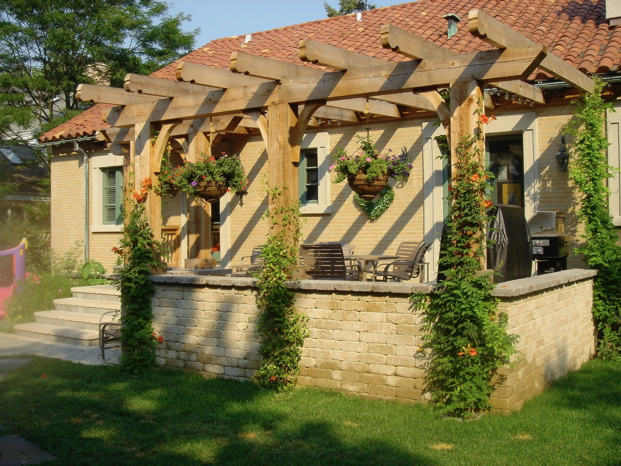 A house with a red tiled roof and brick exterior walls, with a decorative wooden pergola structure on the patio, hanging flower baskets, and climbing plants on the walls. There's a stone retaining wall and outdoor furniture on the patio, with green g