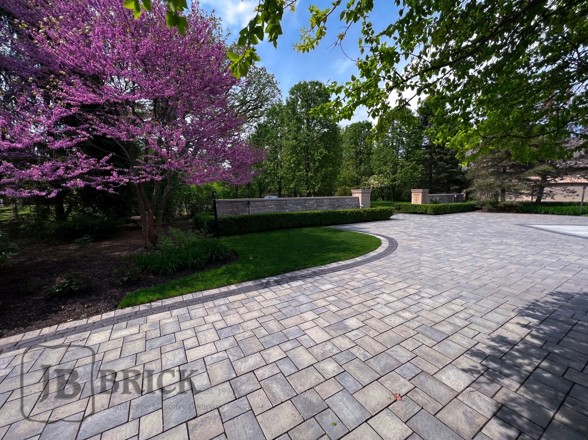 A paved driveway leading to a landscaped backyard with a stone wall, trees with green and purple flowers, and a bright blue sky.