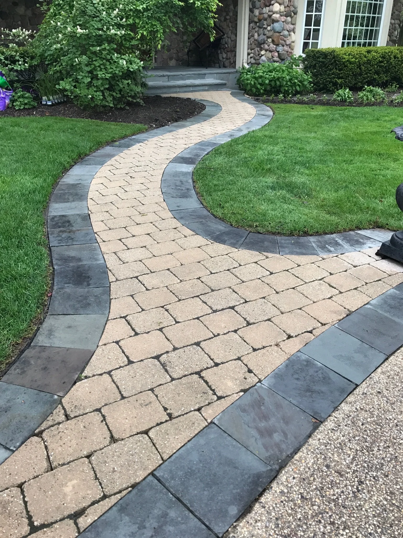 A winding patio pathway made of beige pavers with a gray stone border, surrounded by a well-maintained green lawn and garden plants near a house with stone and siding exterior.
