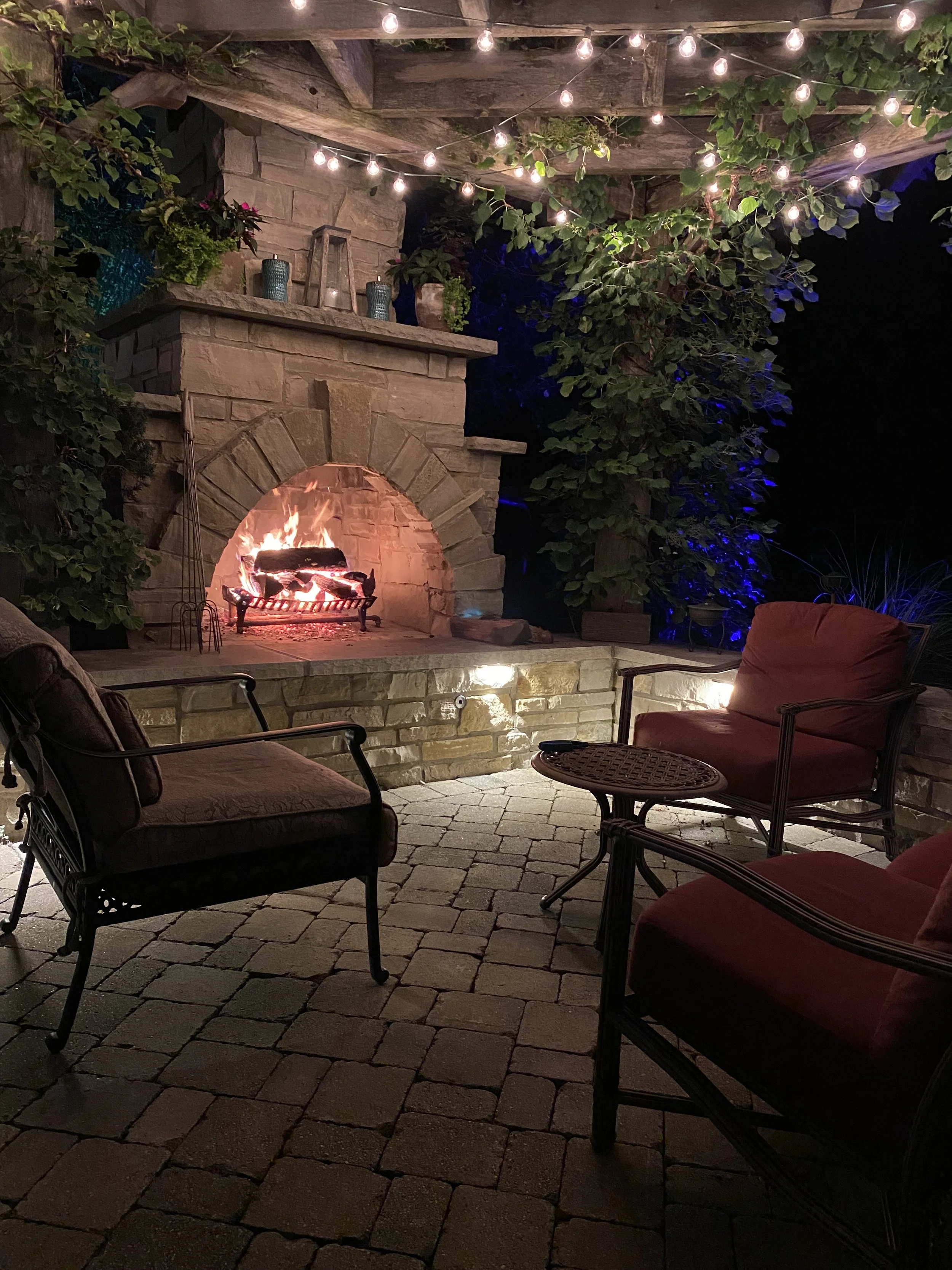 Nighttime outdoor patio scene with a stone fireplace, lit with a fire, surrounded by cushioned chairs, and string lights hanging above. Green plants and foliage are visible around the area.