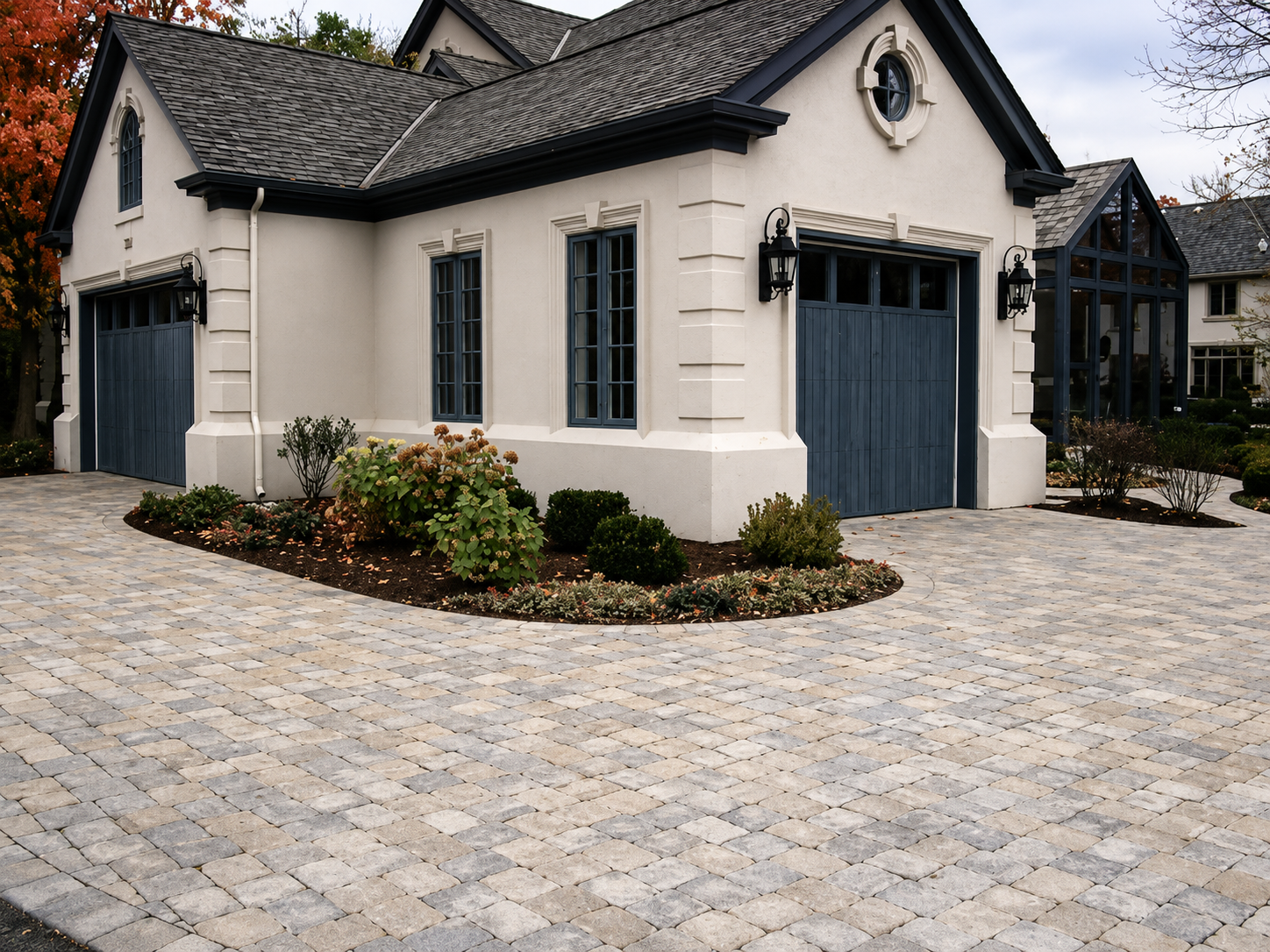 A modern house with a white exterior, dark blue garage doors, and a glass sunroom on the right side. There are black lantern-style lights beside the garage doors, and a landscaped garden with bushes and flowering plants in front. The driveway is pave