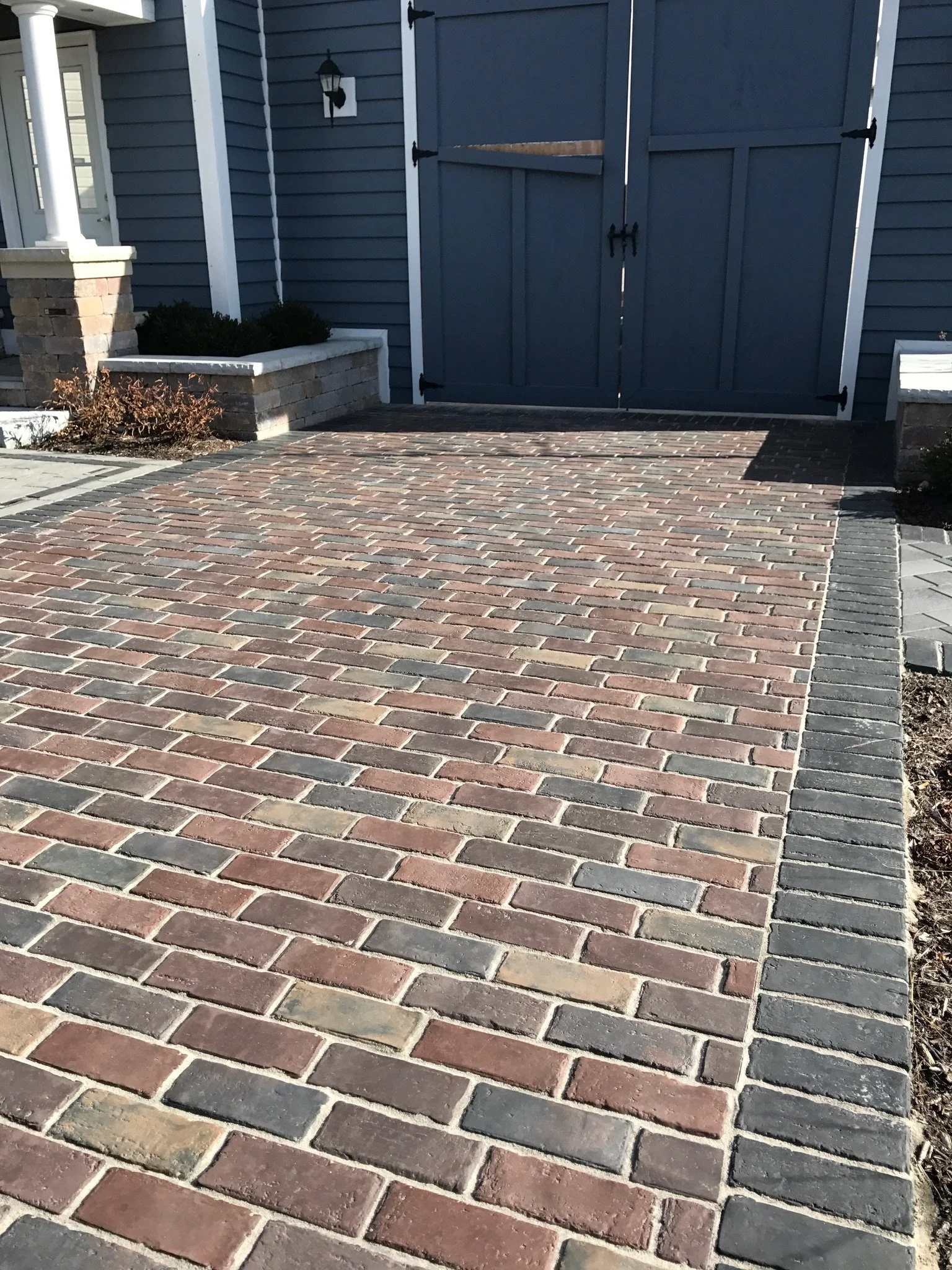 View of a paved brick driveway leading to a closed blue double gate with black hardware, flush against a house with gray siding, white trim, and some shrubbery in a rectangular brick planter.