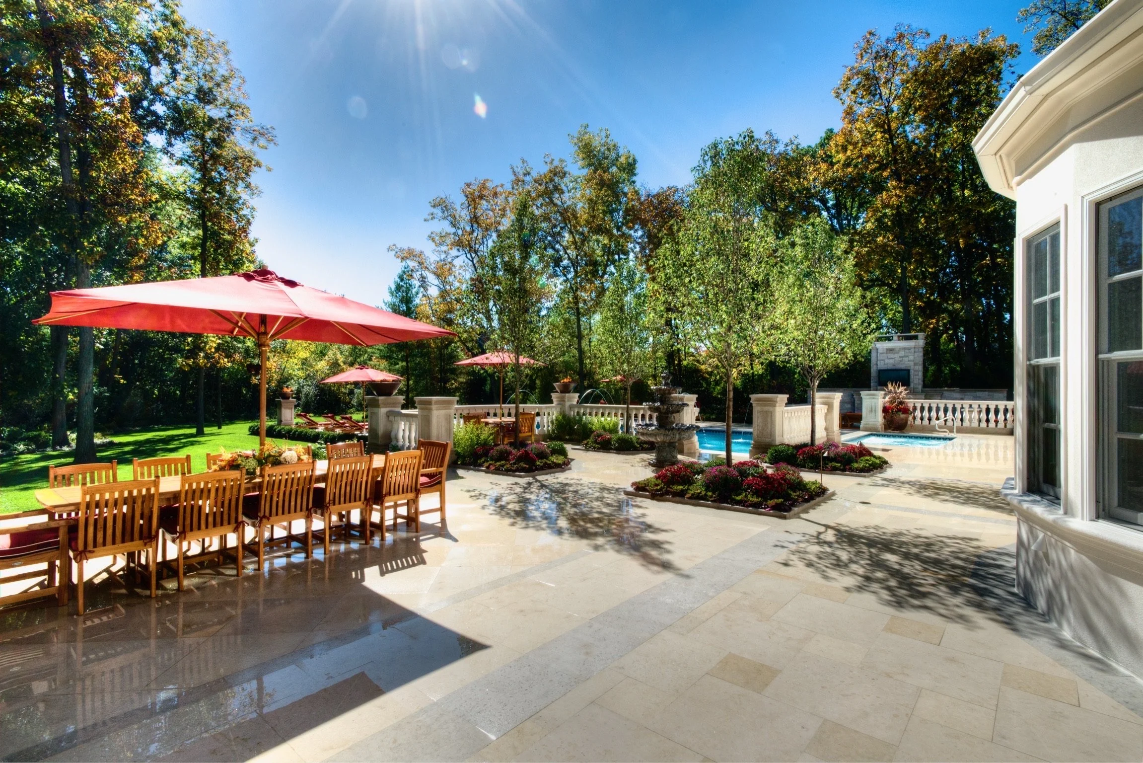 A backyard patio with a large dining table and chairs, red umbrellas, surrounded by lush greenery and trees, with a pool, potted plants, and a house exterior visible under a bright blue sky.