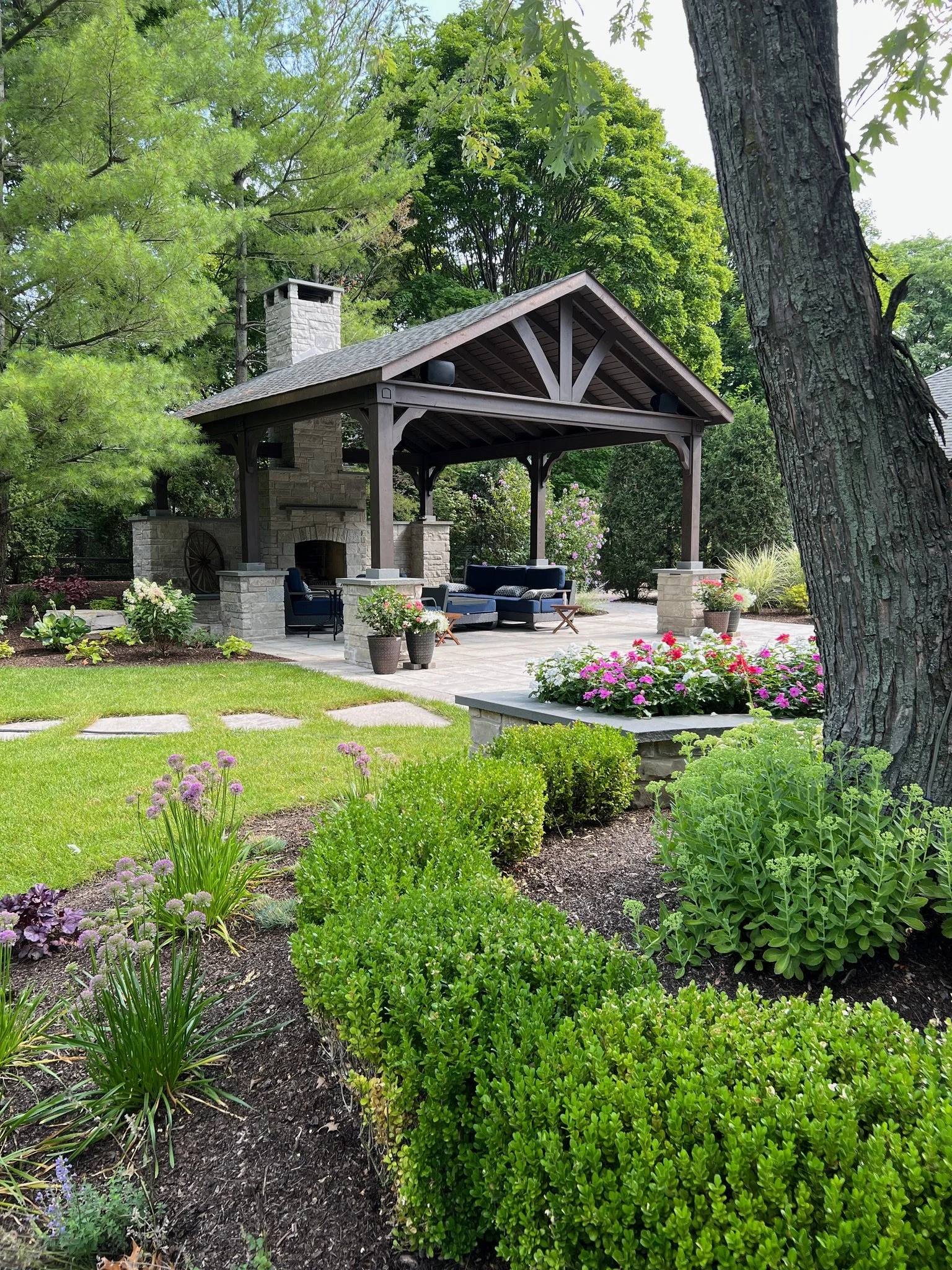 A backyard garden with a stone fireplace and a covered patio area. There are lush green trees, shrubs, and flowering plants surrounding the space, along with outdoor seating.