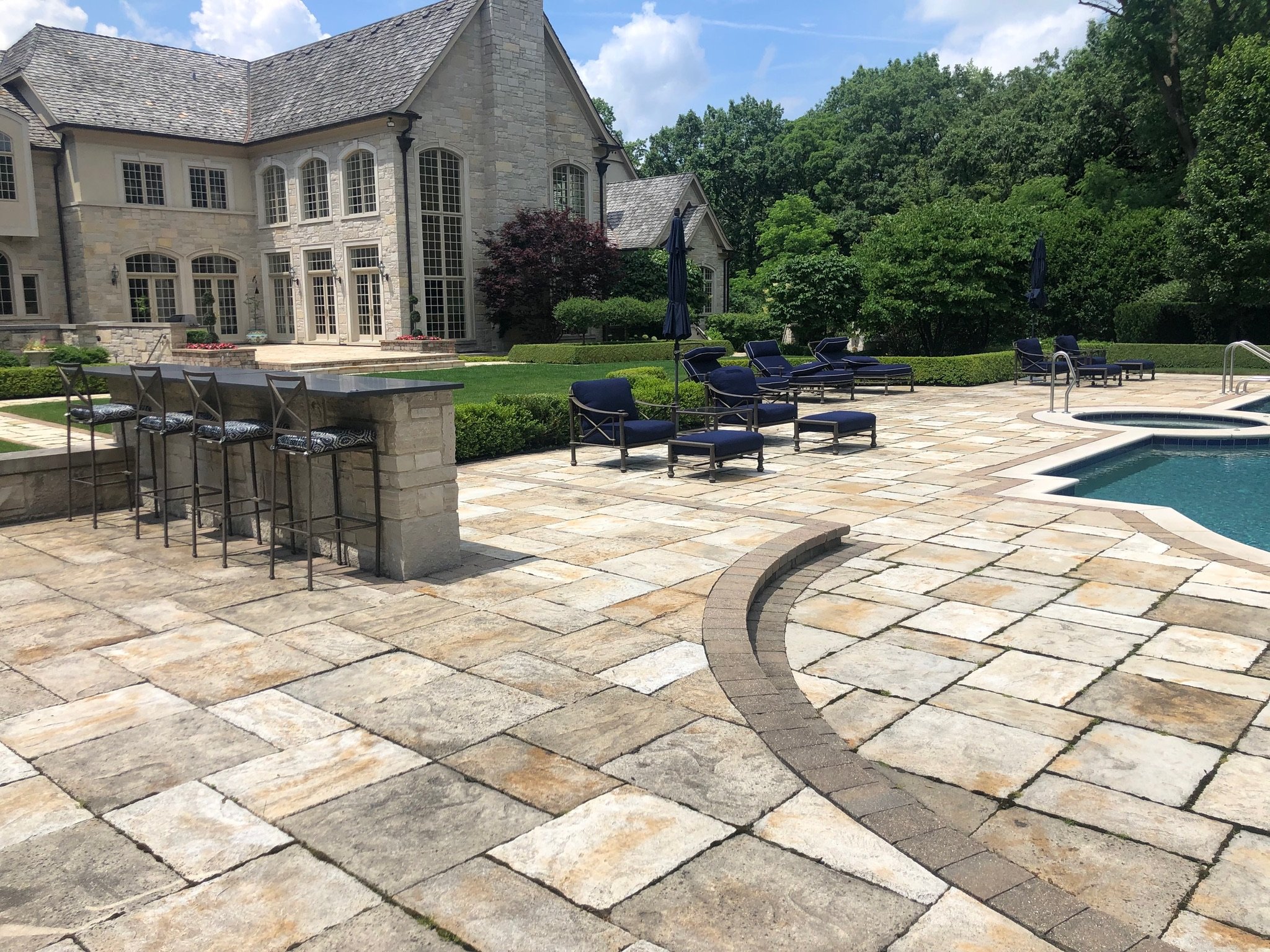 A backyard with a pool, lounge chairs, umbrellas, and a stone patio in front of a large stone house surrounded by greenery and trees.