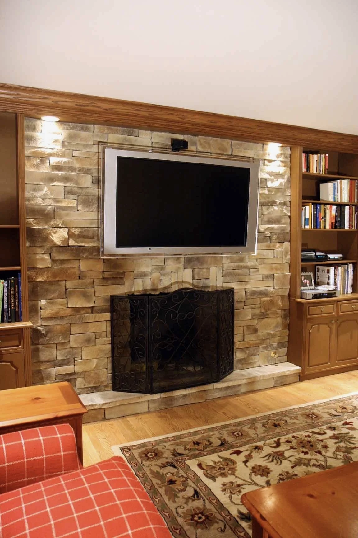 Living room with a stone fireplace, a black screen TV mounted above, wooden built-in bookshelves on both sides filled with books, and a decorative fire screen in front of the fireplace. Part of a wooden dining table and a red checkered cushioned chai
