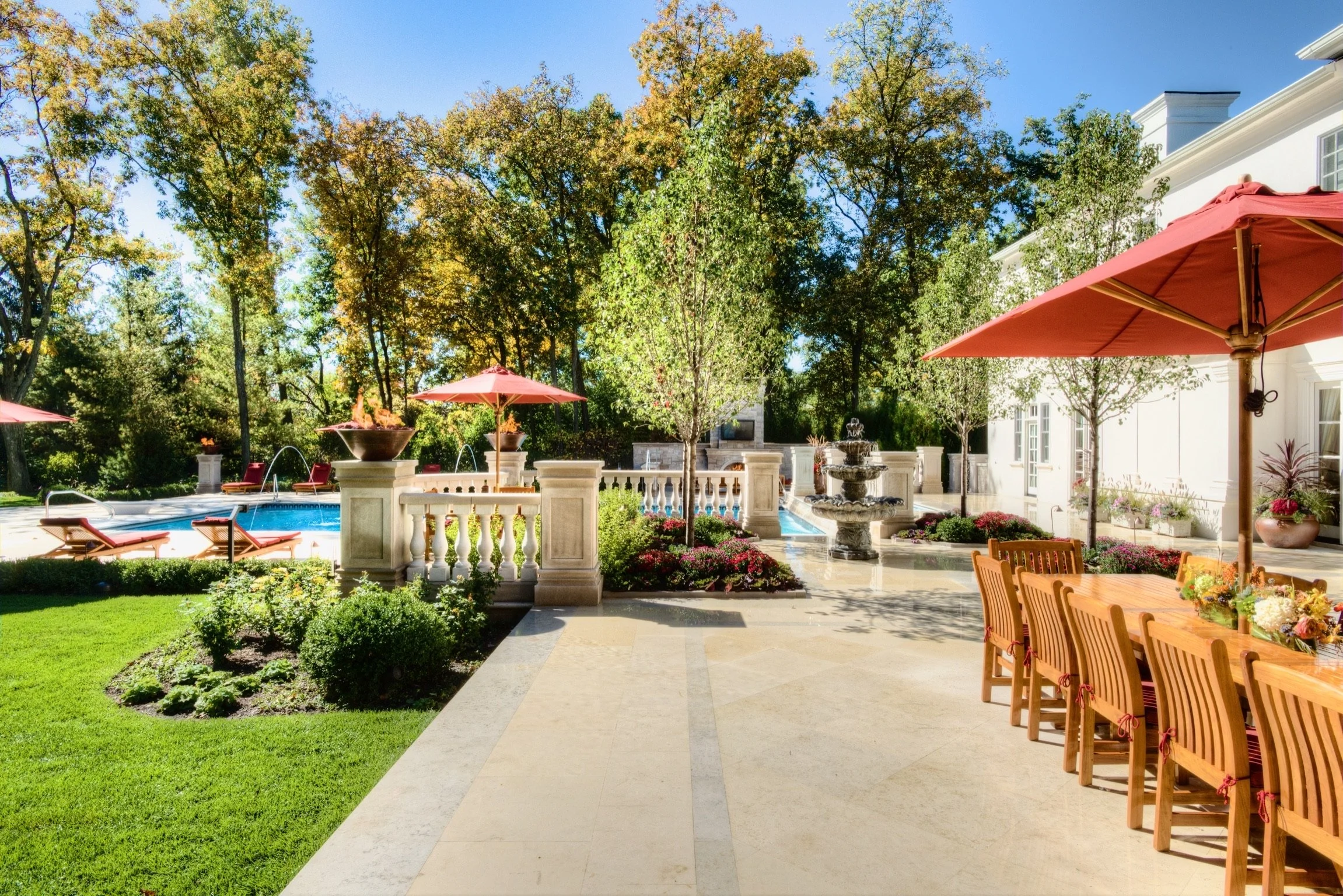 A backyard patio area with outdoor furniture, large umbrellas, a swimming pool, trees with fall foliage, stone fountains, and a white house in the background.
