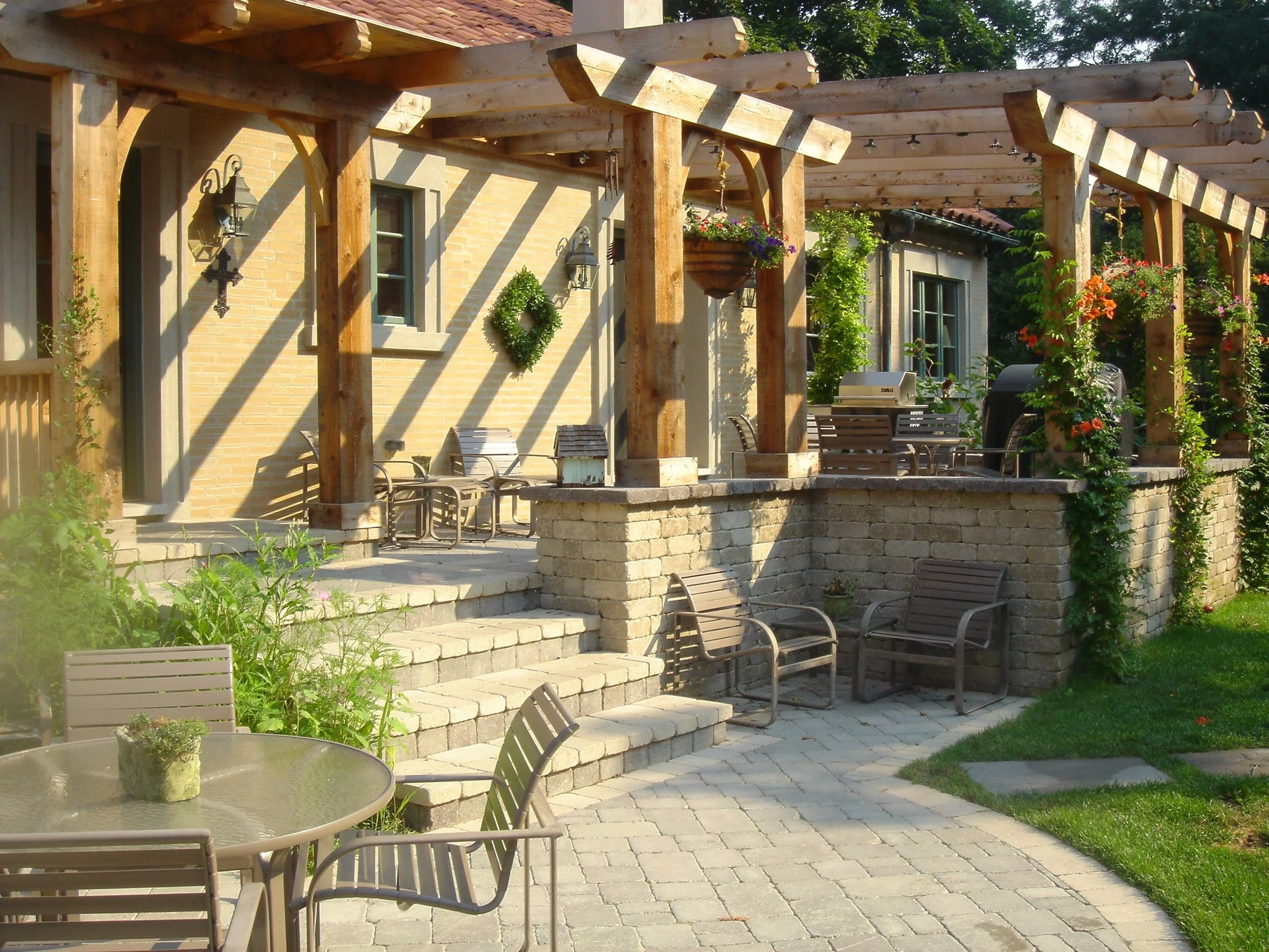 A backyard patio with a round glass table and metal chairs, surrounded by a raised brick patio and lush greenery. There is a wooden pergola with hanging flower baskets and house windows on the house in the background.