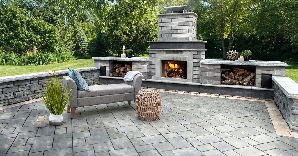Outdoor patio area with a stone fireplace, a gray bench with turquoise cushions, a woven side table, potted plants, and decorative objects on the stone ledges, surrounded by trees and greenery.