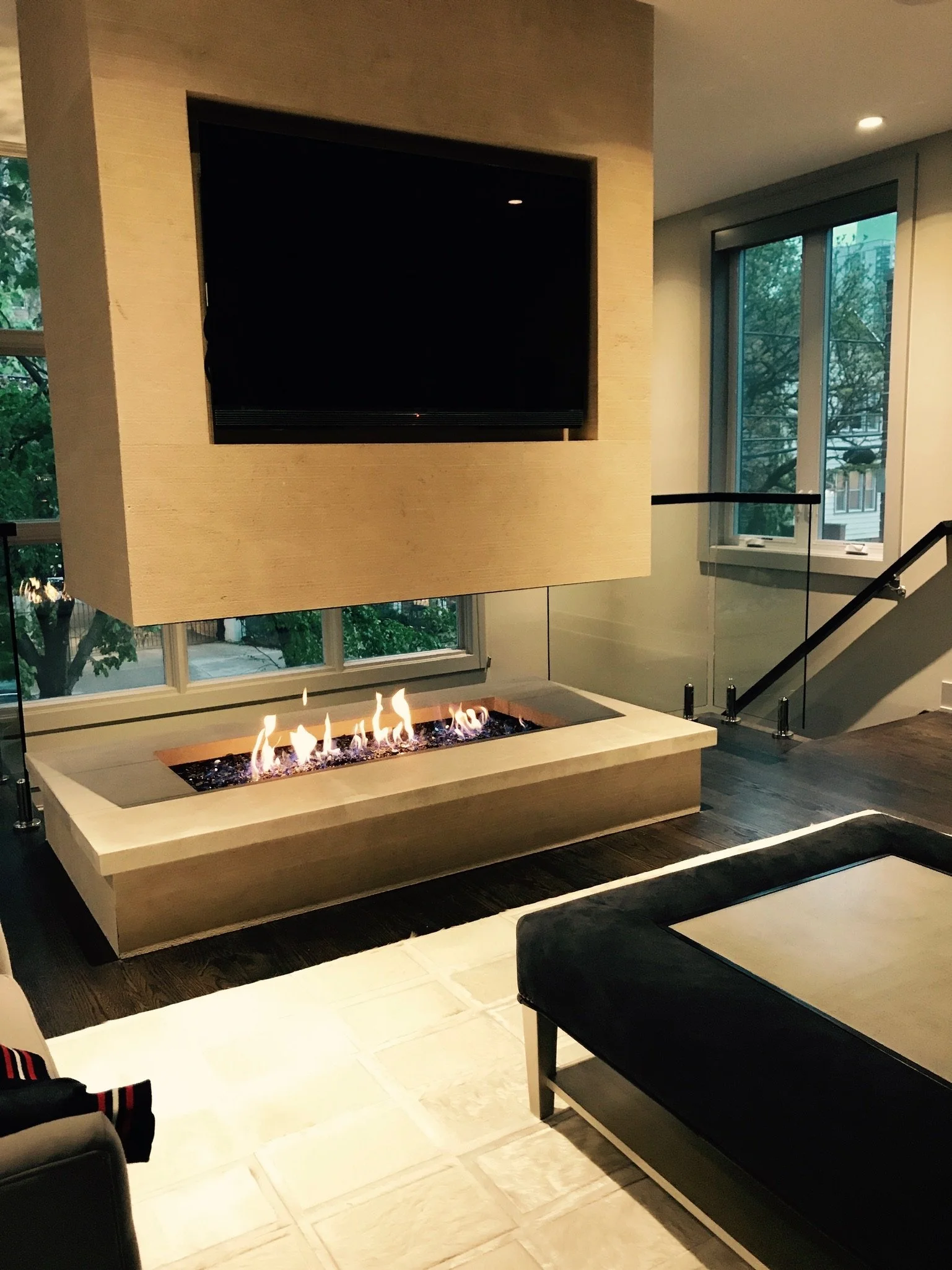 Modern living room with a wall-mounted flat-screen TV, a fireplace with flames, large windows showing trees outside, and black furniture, all illuminated by ceiling lights.