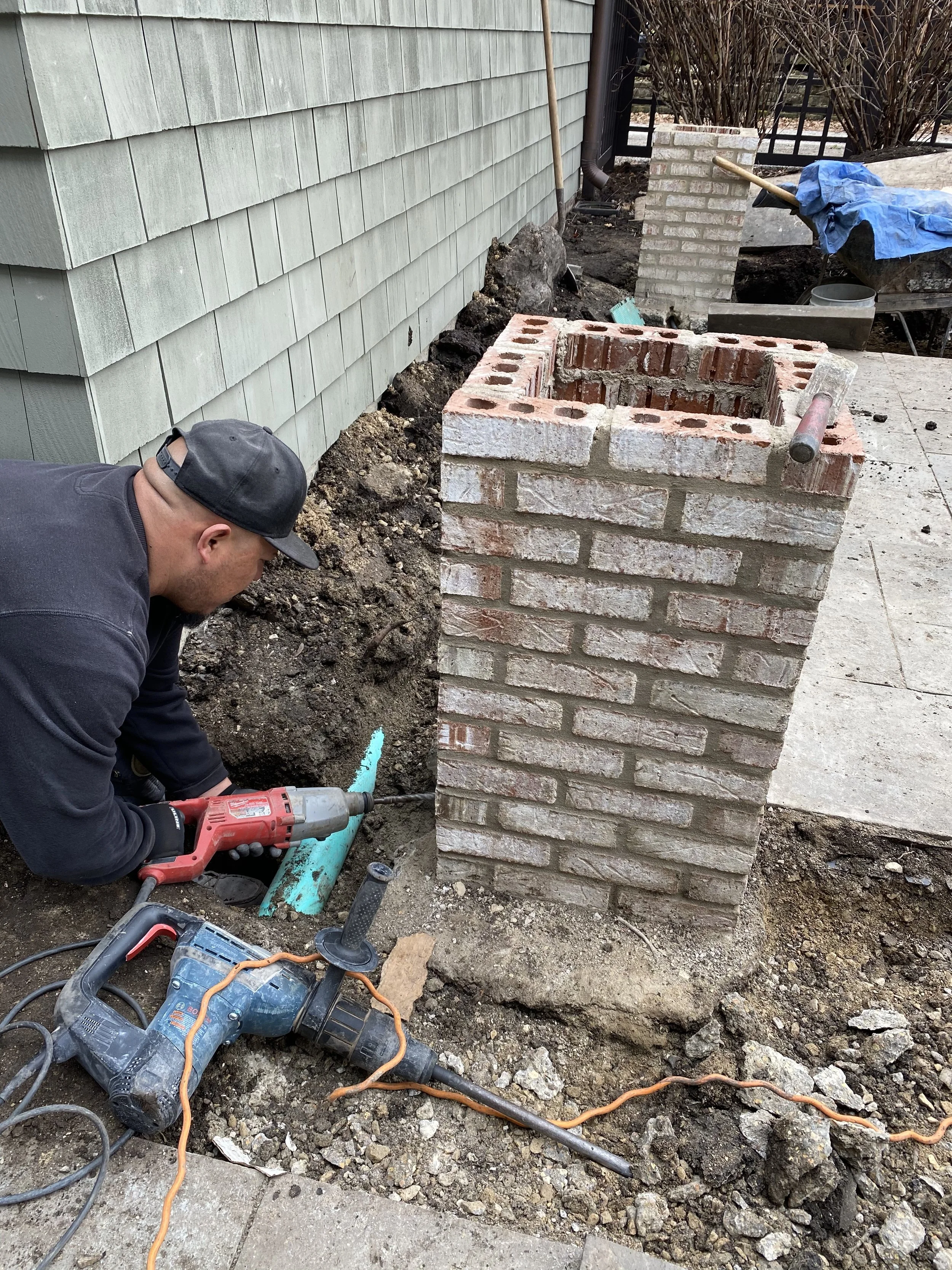 A person working on a construction site with bricks, pipes, and power tools, digging in dirt next to a house with green siding.