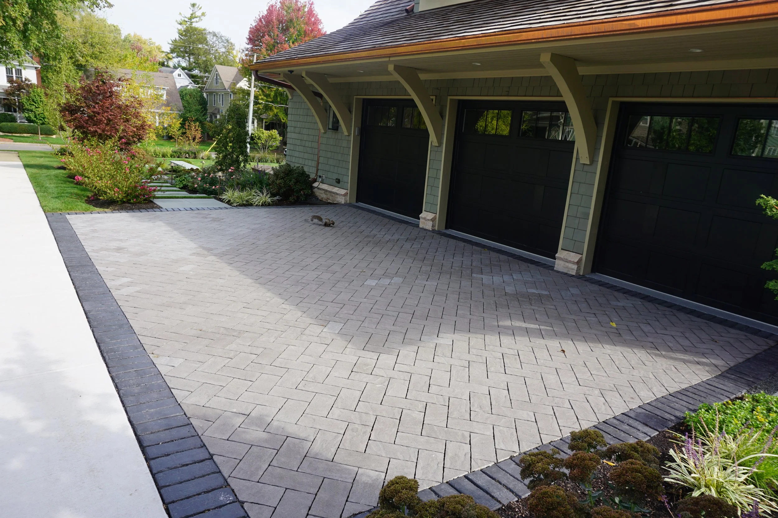 View of a residential garage with black doors, a decorative paver driveway, landscaped flower beds, trees, and neighboring houses in the background.