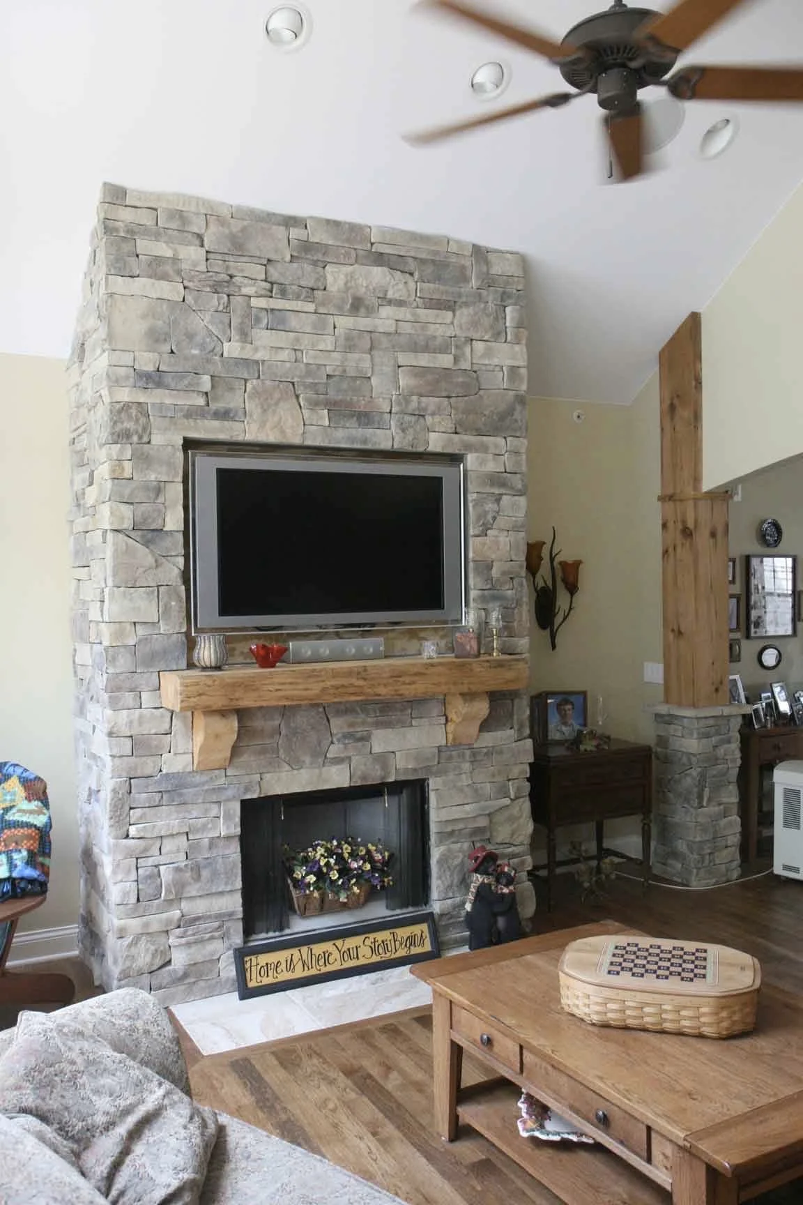 Living room with stone fireplace, wall-mounted TV, wooden mantel, ceiling fan, and framed photos.