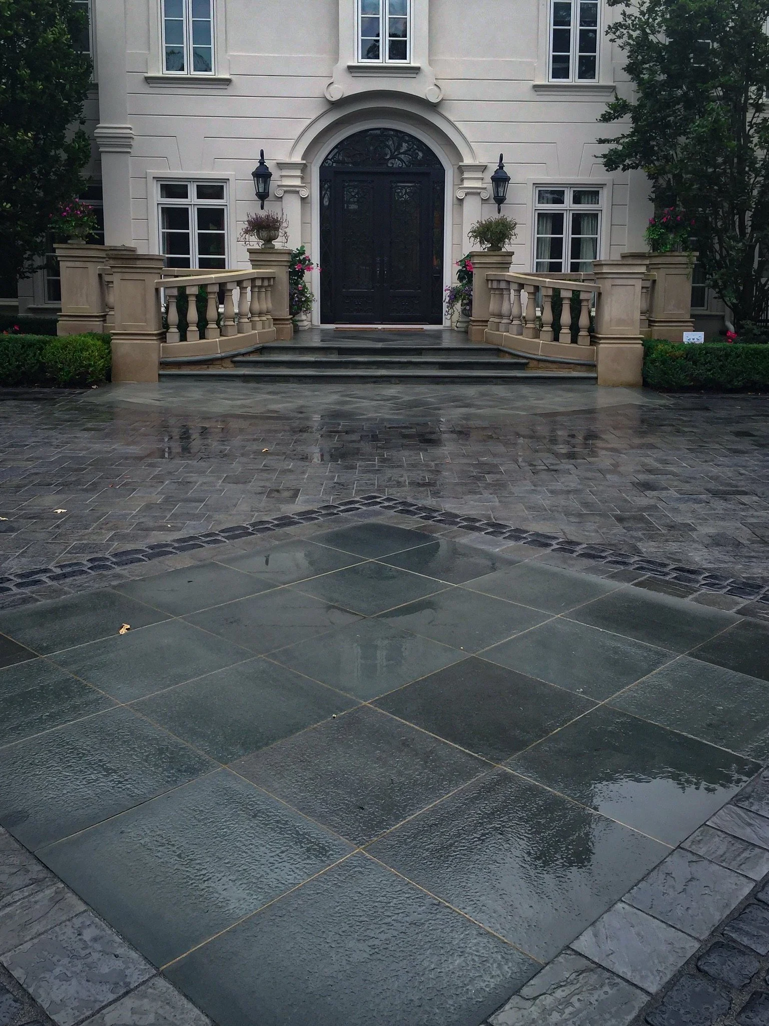Front view of a large, elegant house with a black front door, steps leading up to the door with a beige railing, surrounded by potted plants, bushes, and trees, with a wet stone walkway in the foreground.