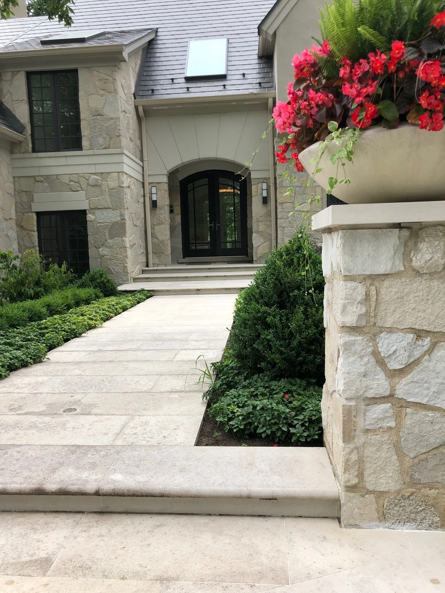 Front view of a house entrance with stone walls, black front door, stairs, stone planters with red and pink flowers, green shrubs, and a pathway.
