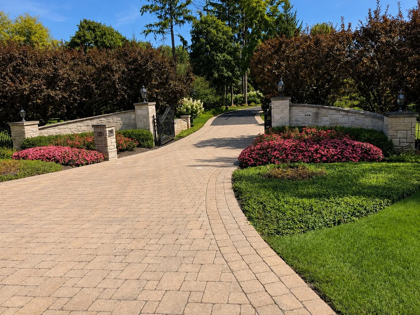 A paved driveway leading to a gated entrance flanked by stone pillars with lamps, surrounded by well-maintained flower beds with pink blossoms and lush green plants, with trees and a blue sky in the background.
