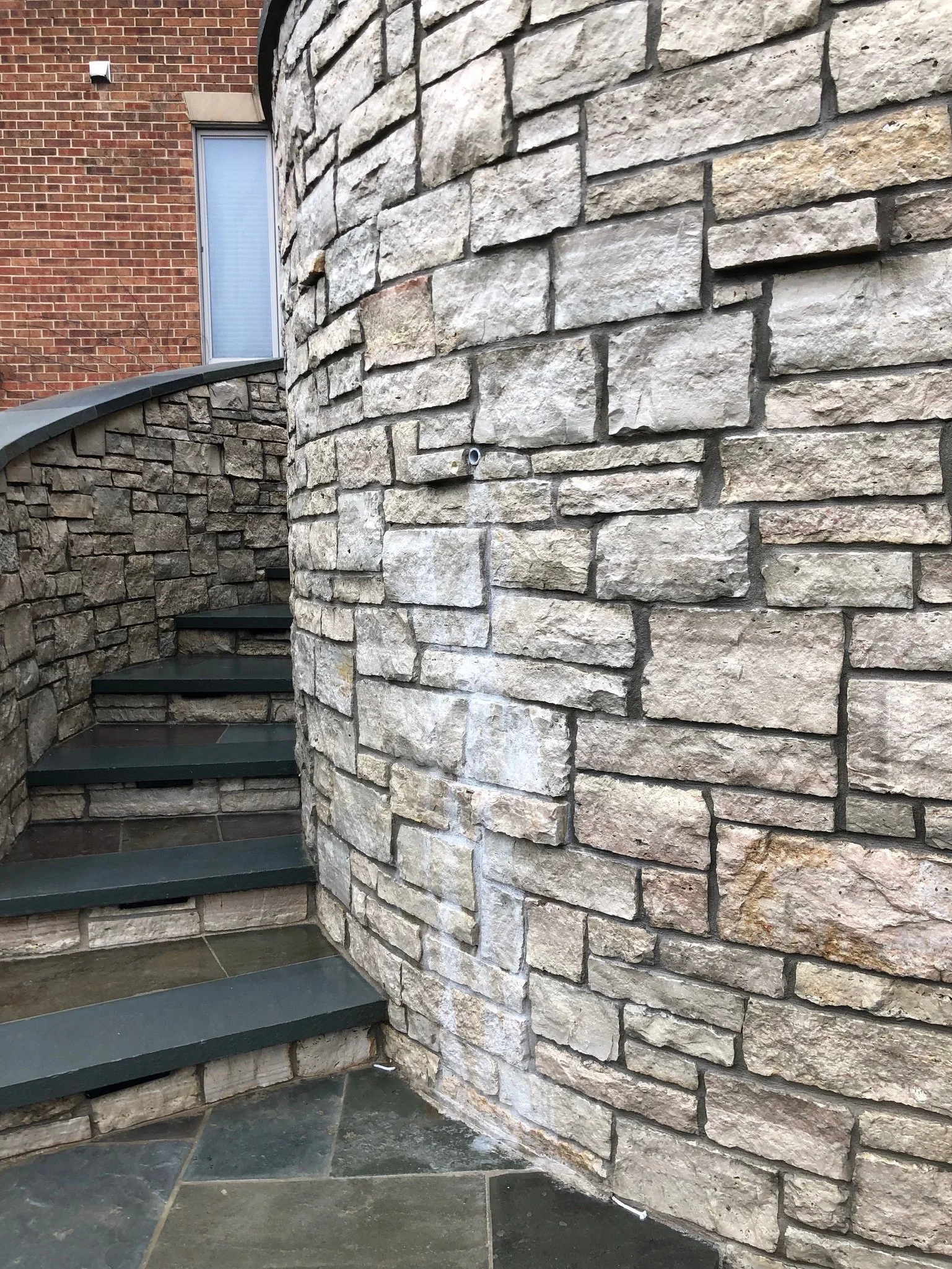 Close-up of a curved stone exterior wall with beige and gray bricks, next to a brick building with a window, and a set of green stairs with a railing.