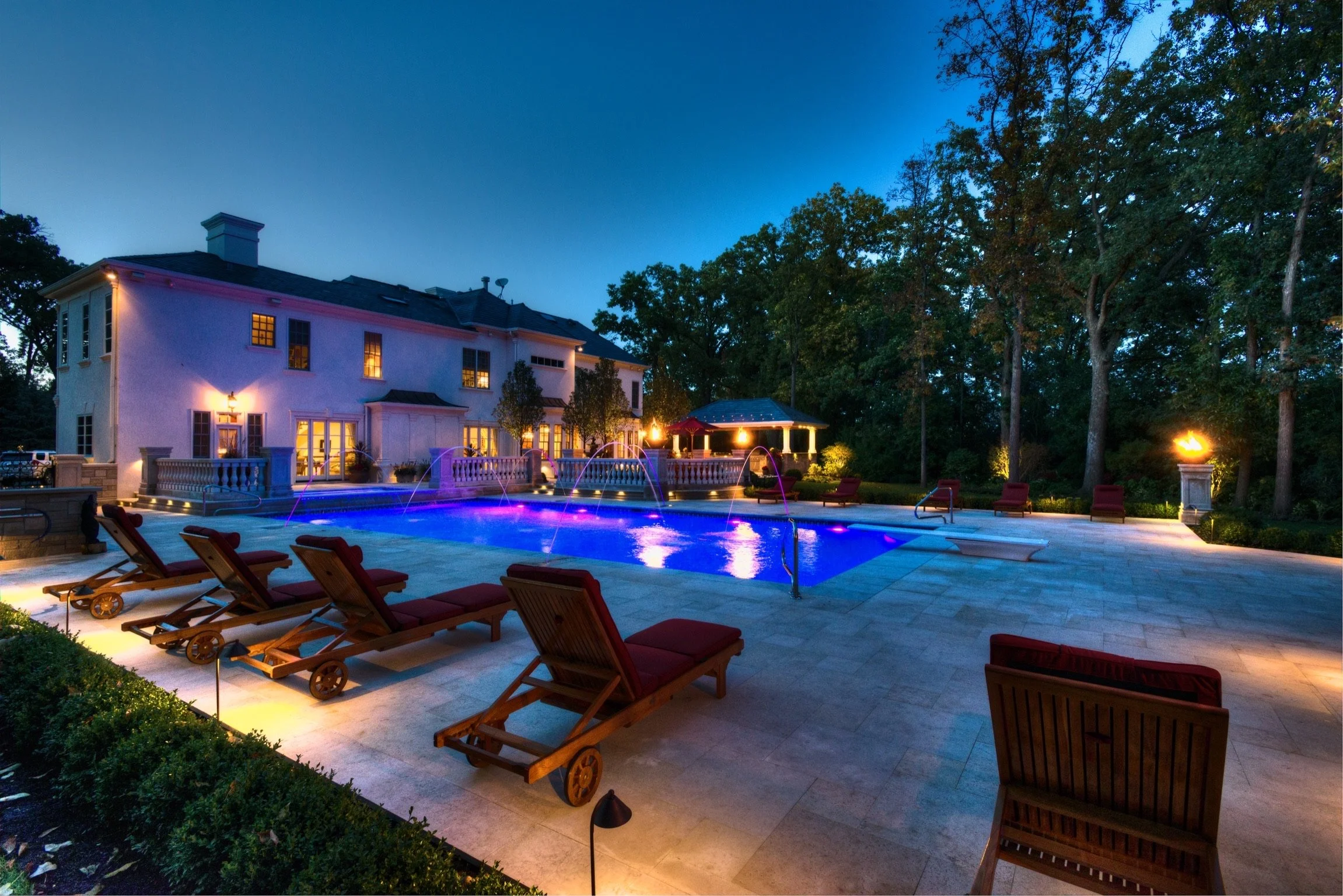 A house with illuminated windows behind a backyard swimming pool with pink and purple lights. Lounge chairs are arranged around the pool, and trees surround the area at dusk or night.
