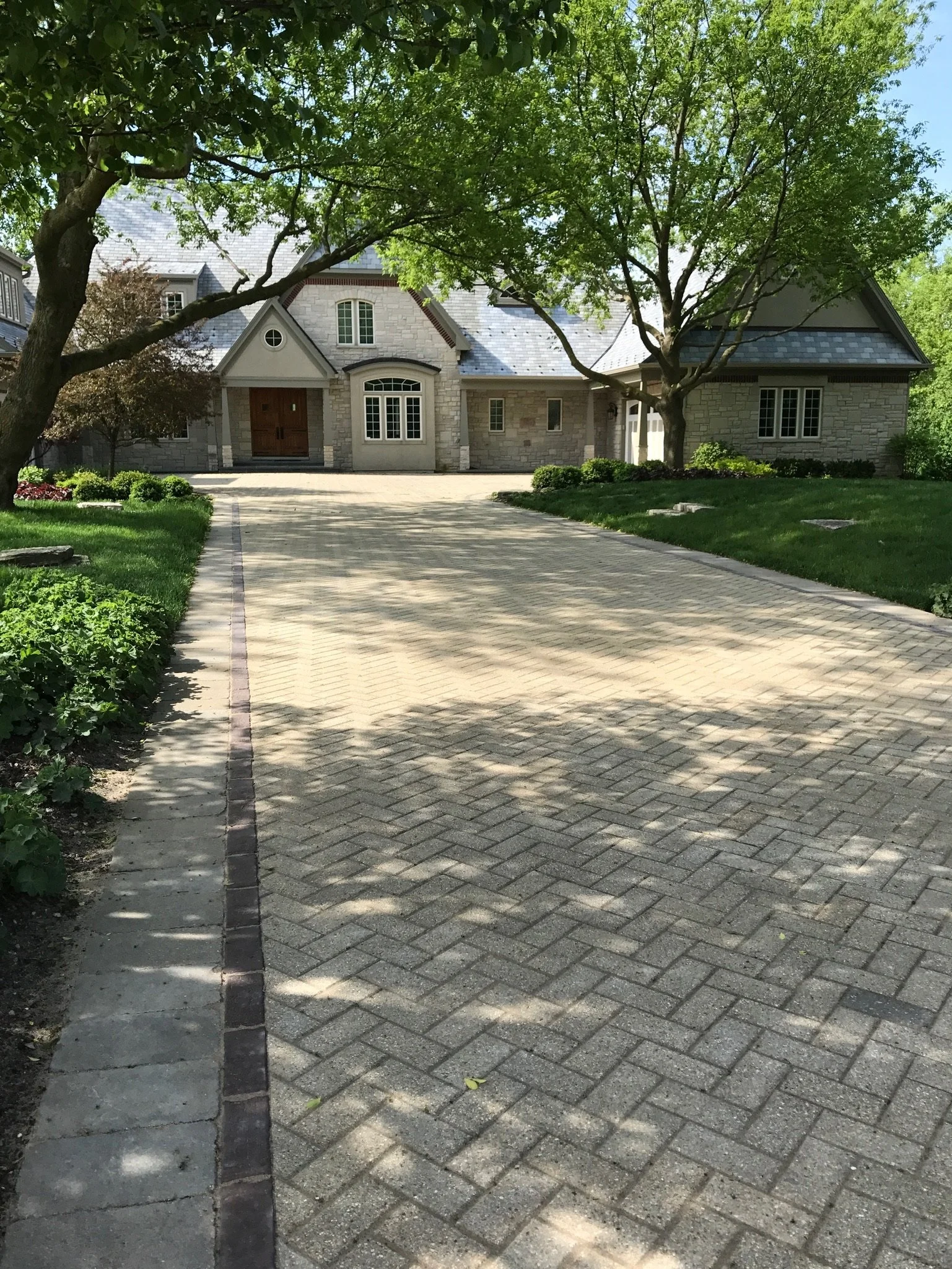 A brick driveway leading up to a house with stone exterior walls and a gabled roof. There are two large trees with green leaves on either side of the driveway, casting shadows on the ground. The house has white framed windows and a wooden front door.