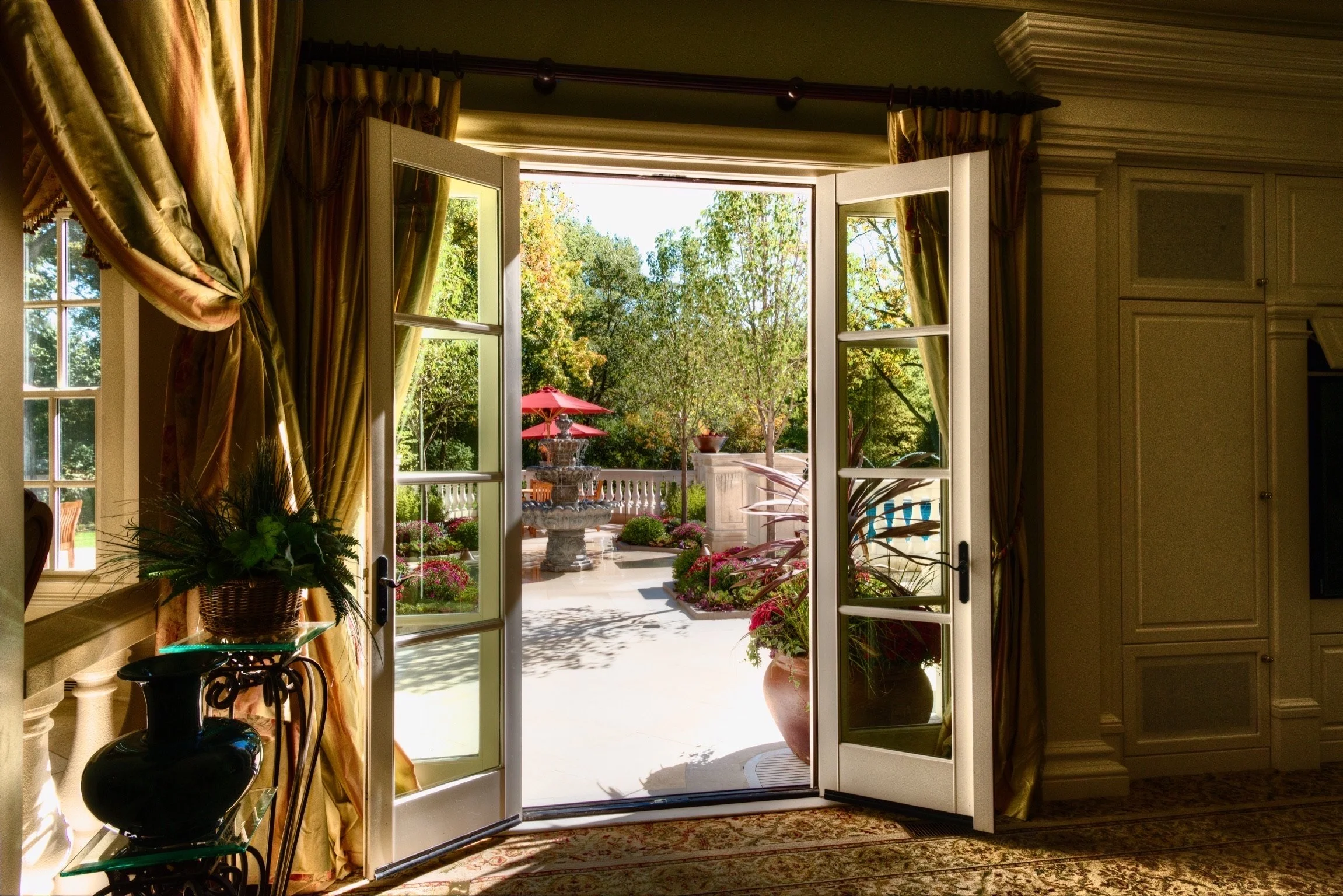 Sunlit French doors open to a patio with a fountain, lush greenery, and a red umbrella.