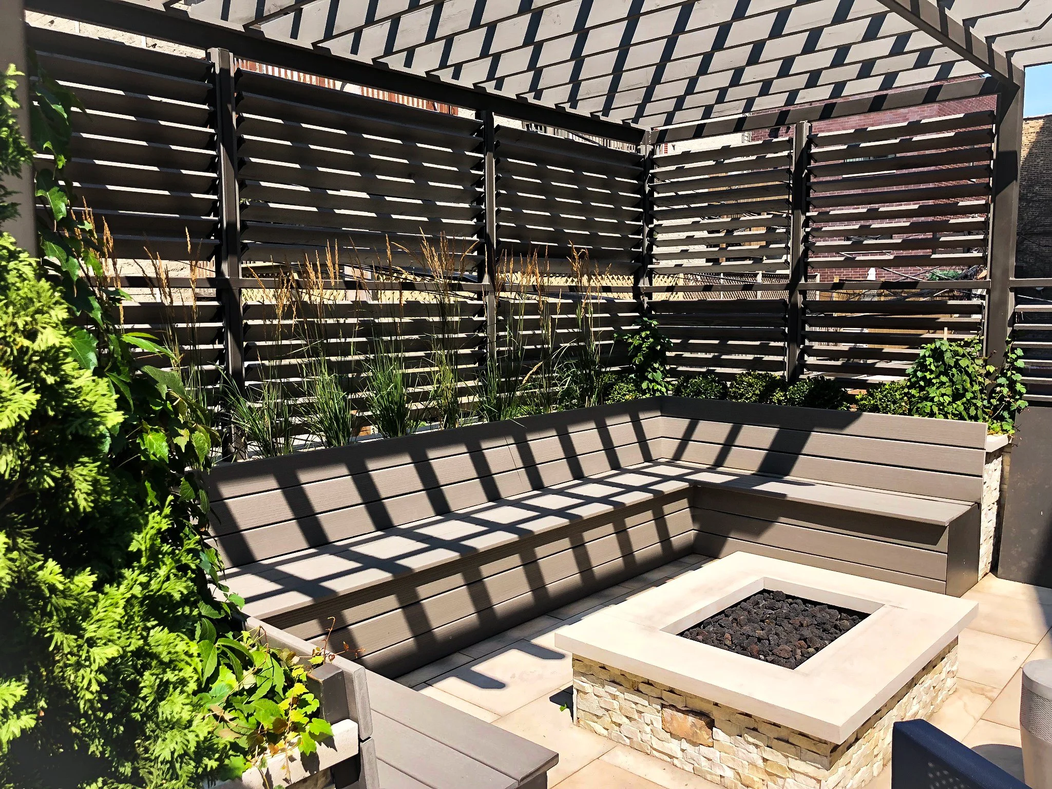 A modern outdoor patio space with a wooden slat privacy fence, a built-in bench, a planter with dark rocks, and various green plants. The space is shaded by a pergola with diagonal slats, creating striped shadows.