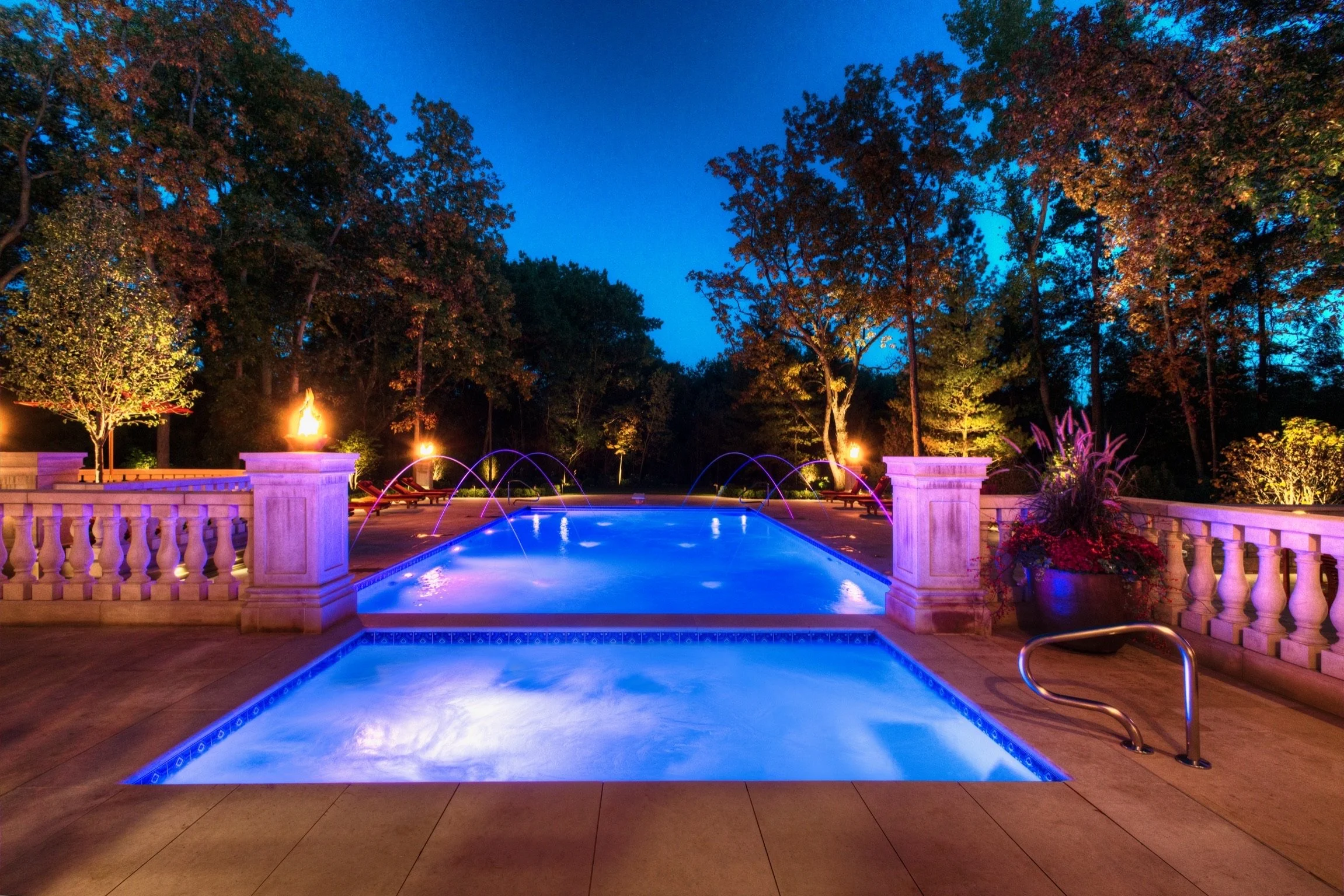 A lit swimming pool at night with blue lighting, surrounded by a stone deck and decorative balustrades. In the background, tall trees are illuminated by outdoor lighting, and a fire feature adds warmth and ambiance. The sky is dark blue, indicating e