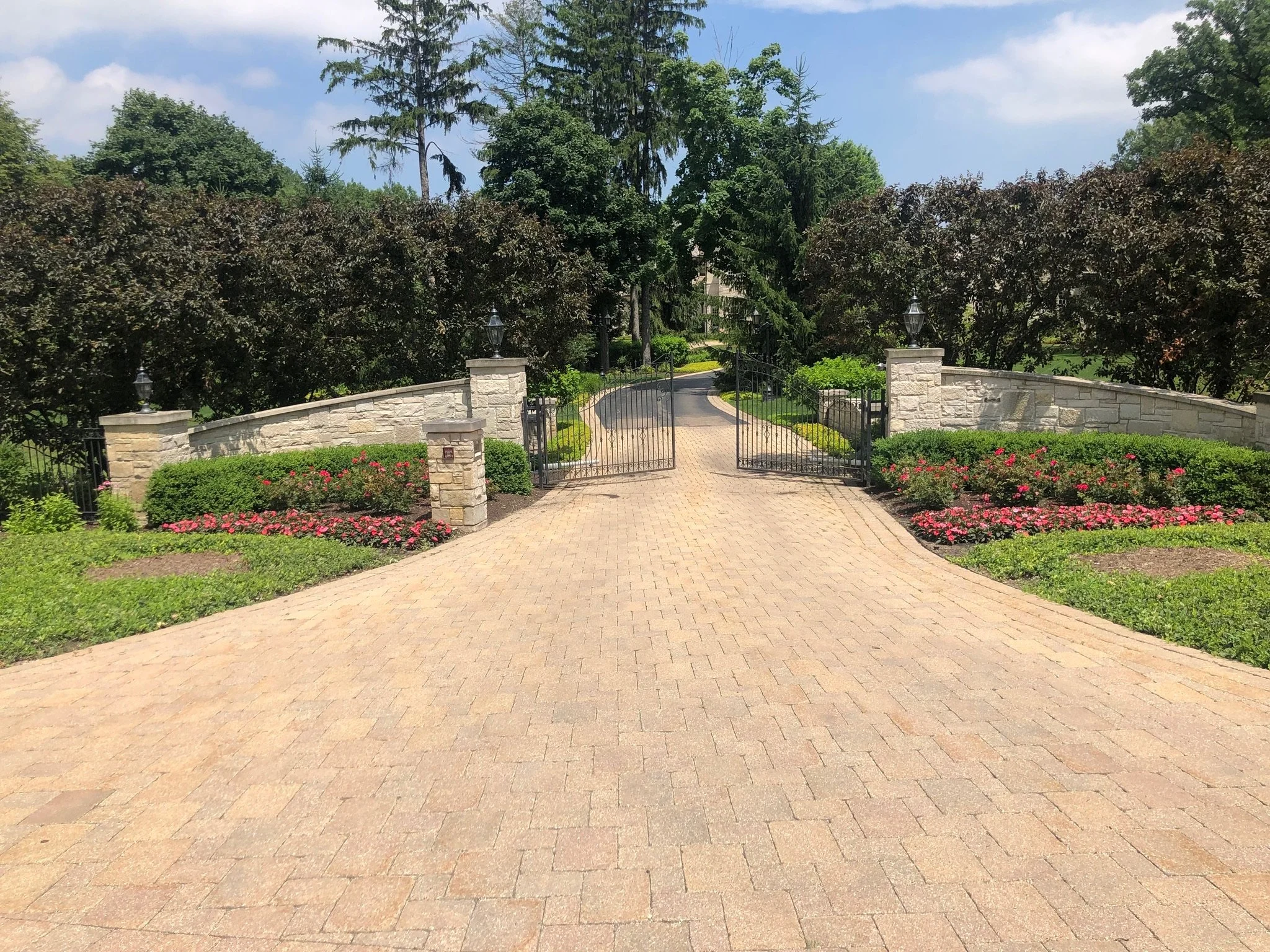 A paved brick driveway leading to a gated entrance with stone columns and black metal gates, surrounded by well-kept garden beds with pink flowers and green bushes, and tall trees in the background under a partly cloudy sky.