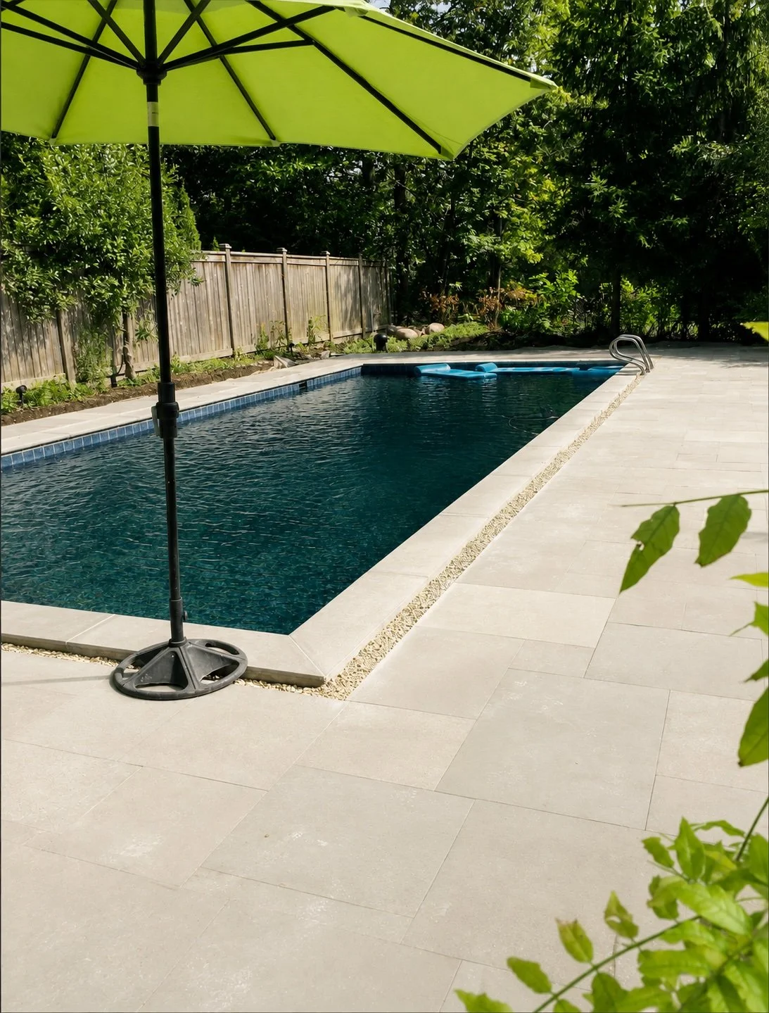 A backyard swimming pool with a green patio umbrella on the side, surrounded by trees and a wooden fence.