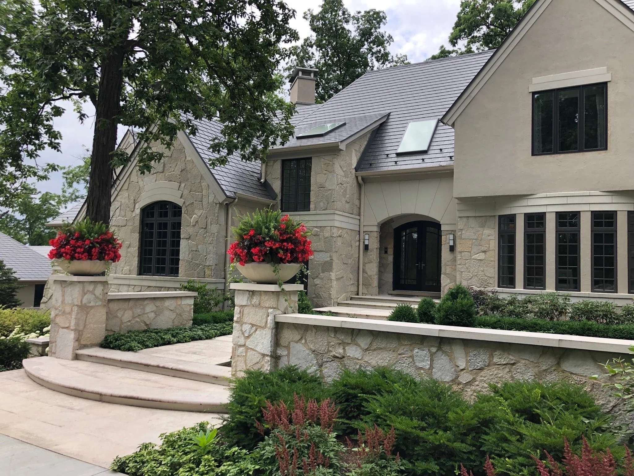 A large, elegant house with a stone facade, black window frames, and a gray tiled roof, surrounded by a landscaped garden with potted plants and bushes.