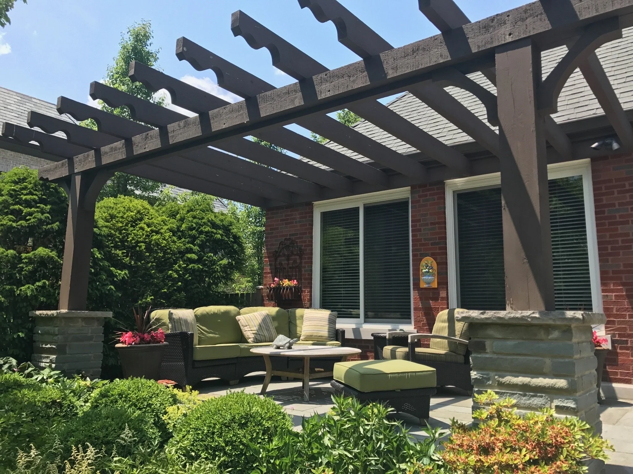 A backyard patio with outdoor furniture including couches and chairs with green cushions, a table, and potted flowers, under a wooden pergola, with a brick house and green trees in the background.
