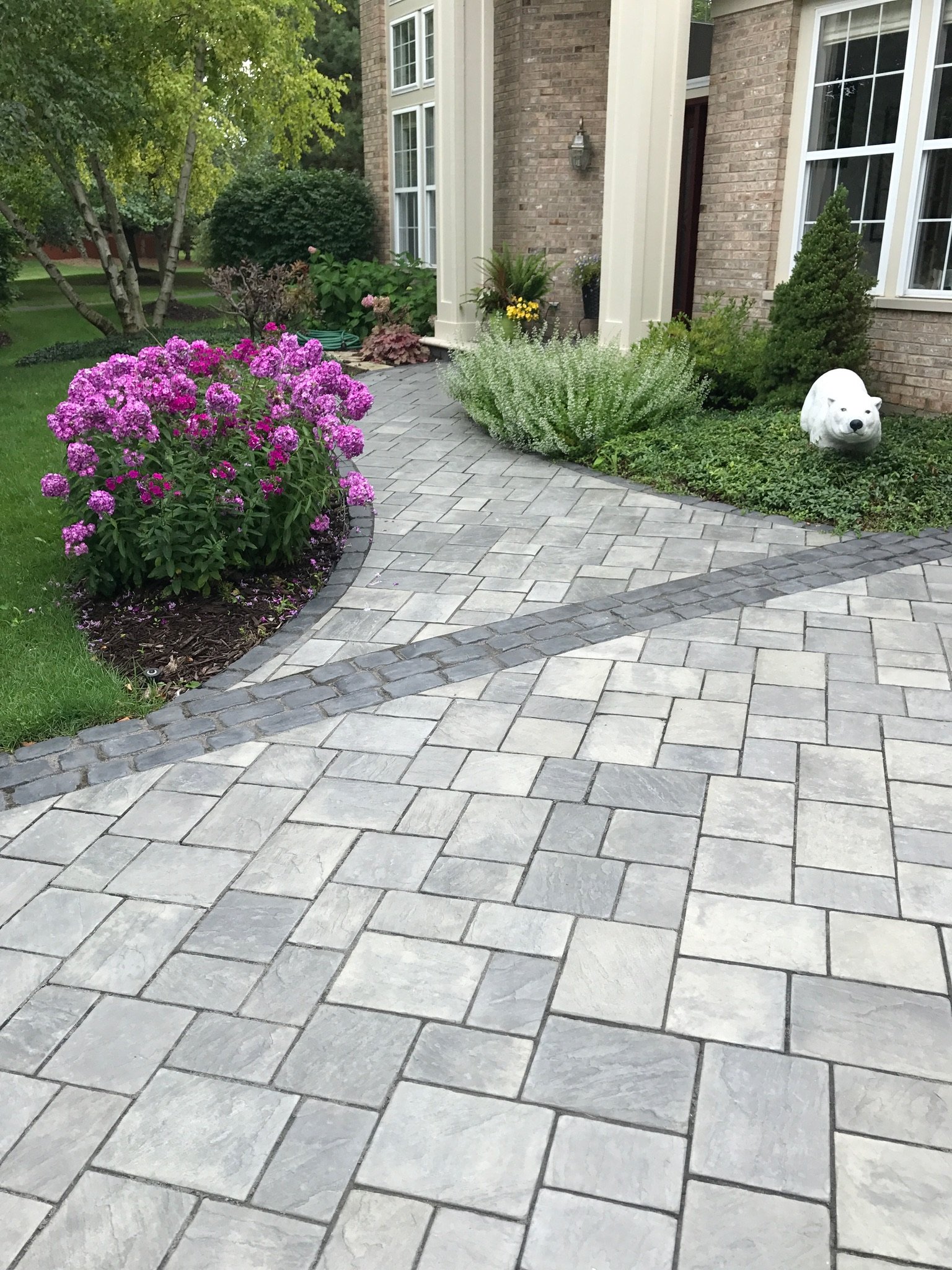 A front yard with a brick house, a paved walkway, pink flowers, shrubs, and a white bear-shaped garden decoration.