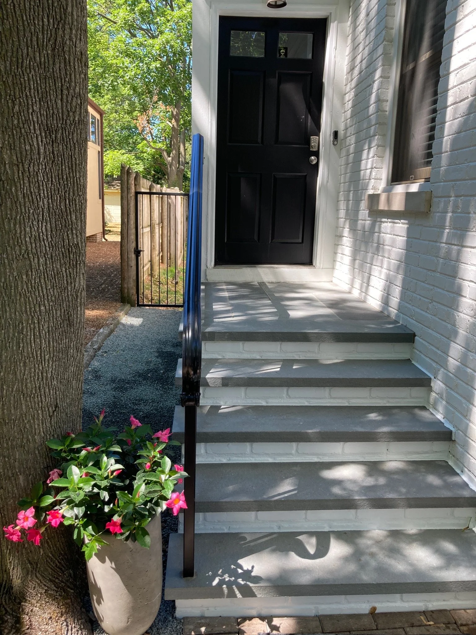 Front porch with black door, white brick exterior, gray stairs, black handrail, potted pink flowers, tree on the left, and green trees in the background.