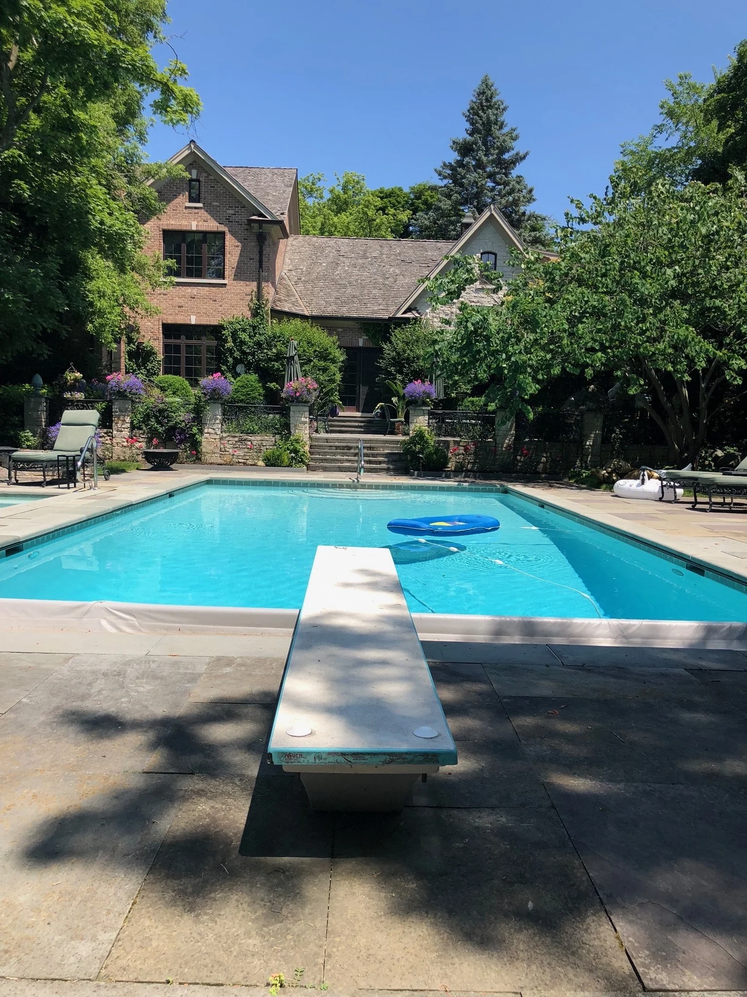 A swimming pool with a diving board, surrounded by patio furniture and flowering plants, overlooking a large house with brick walls and a sloped roof, with trees and a blue sky in the background.