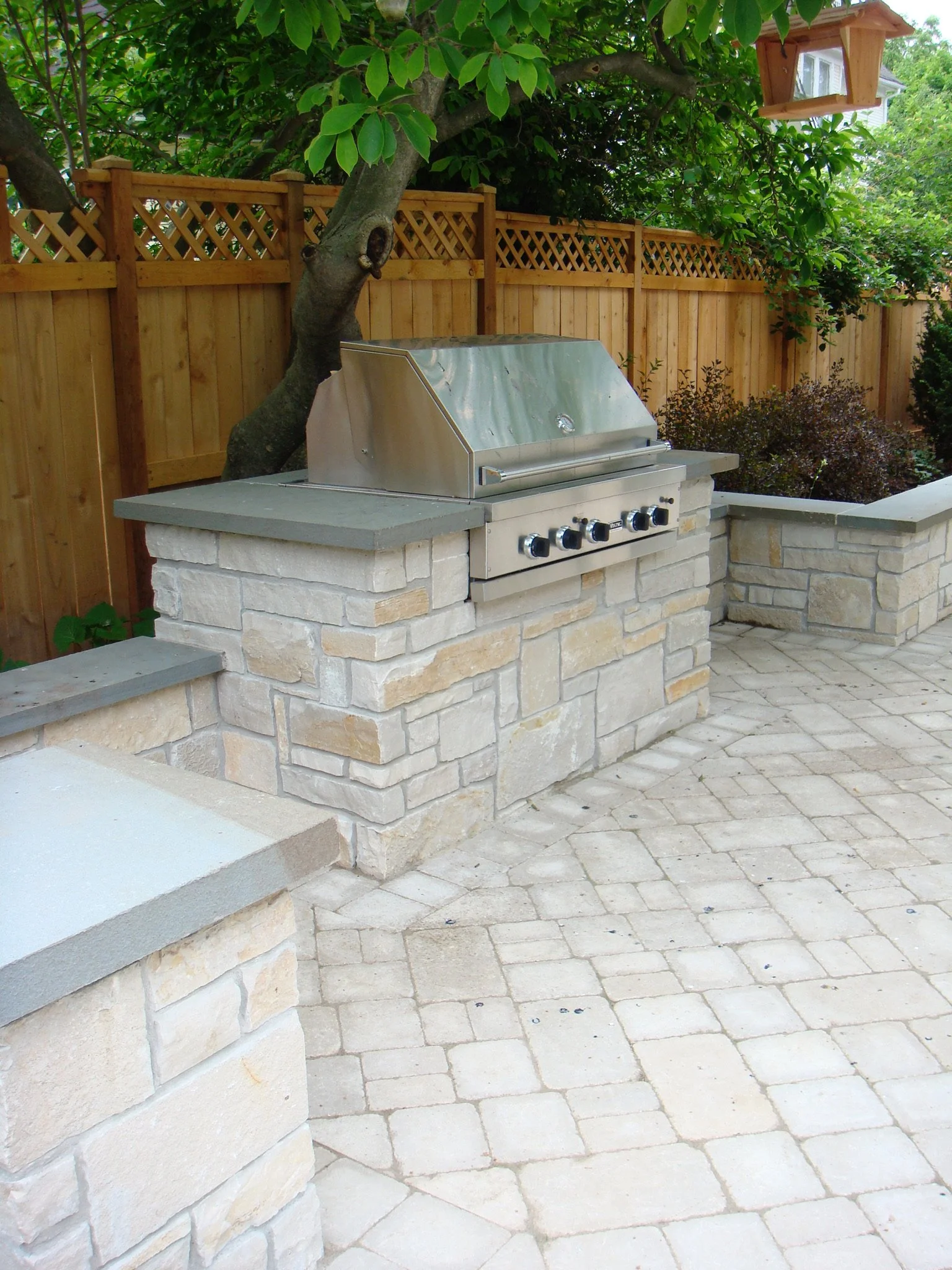 A backyard patio with a stone barbecue grill, stone patio flooring, a wooden fence, and a birdhouse hanging from a tree with green leaves.