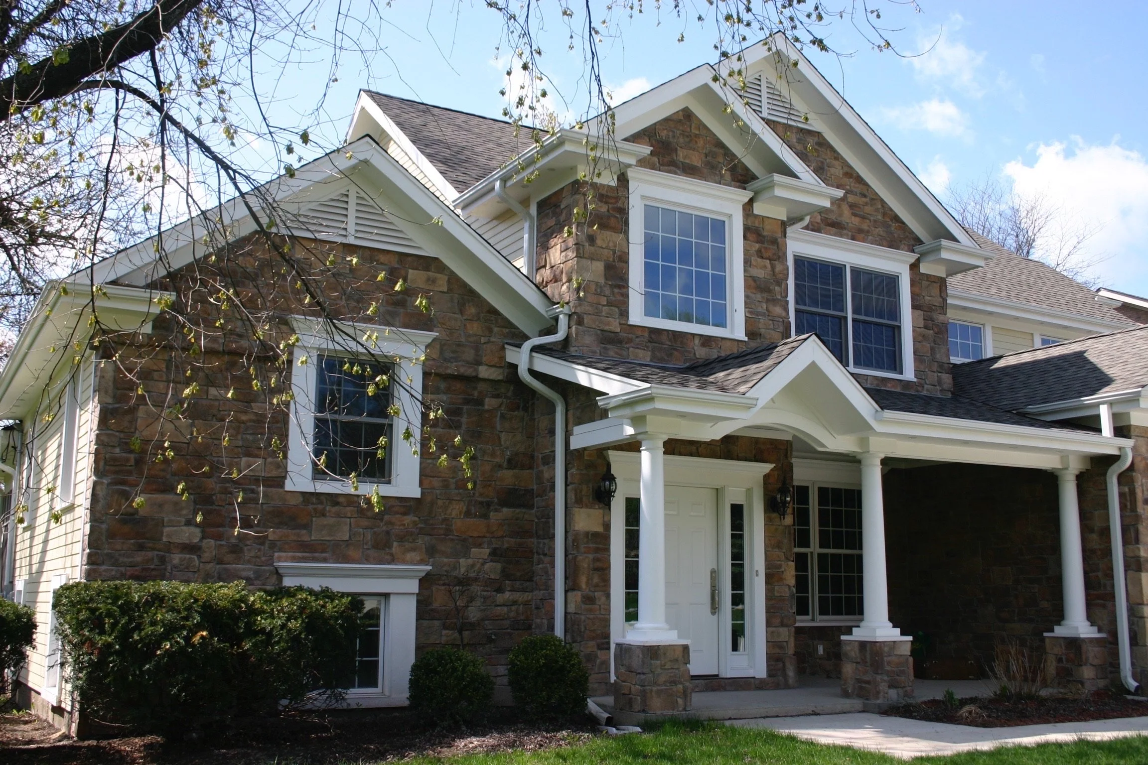 Front view of a two-story house with stone and siding exterior, white trim, multiple large windows, a small porch with columns, and a well-maintained lawn.
