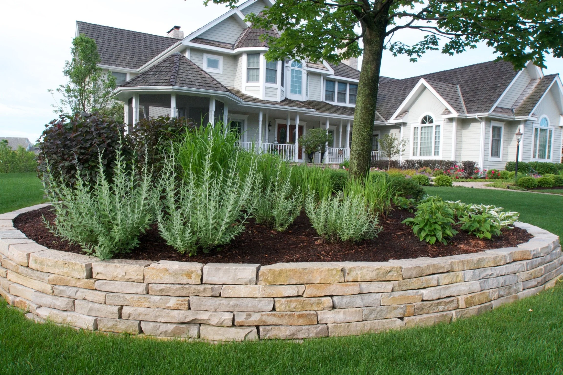 A brick raised garden bed with green plants, surrounded by a well-manicured lawn and a large house with white siding and multiple windows.