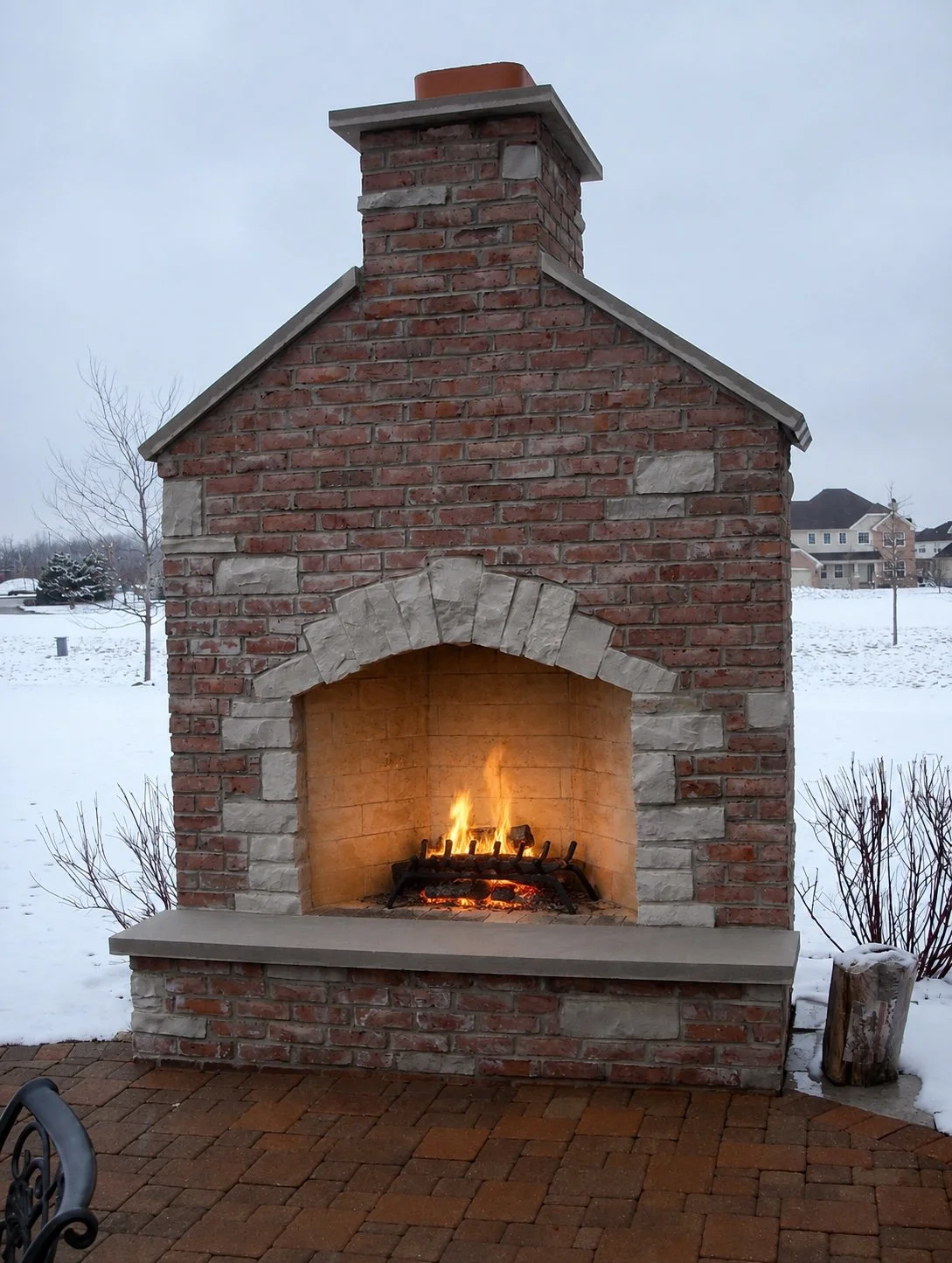 Outdoor brick fireplace with a fire burning inside, set on a brick patio in a snowy landscape.