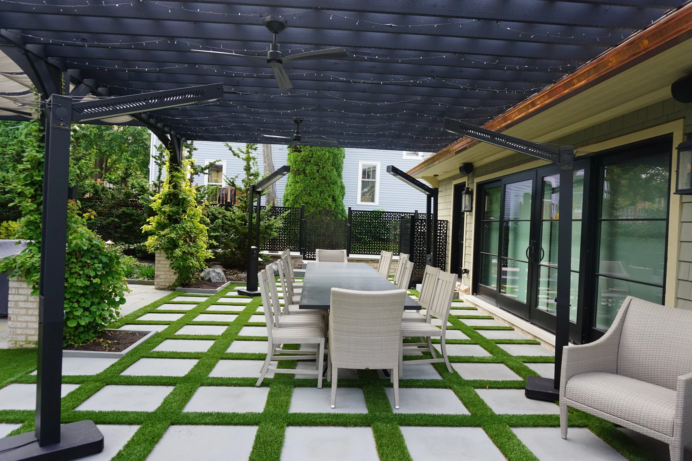 An outdoor patio area with a rectangular dining table and multiple chairs on a decorative lawn with square concrete pavers. The space is covered by a dark blue pergola with string lights and ceiling fans, adjacent to a house with sliding glass doors 