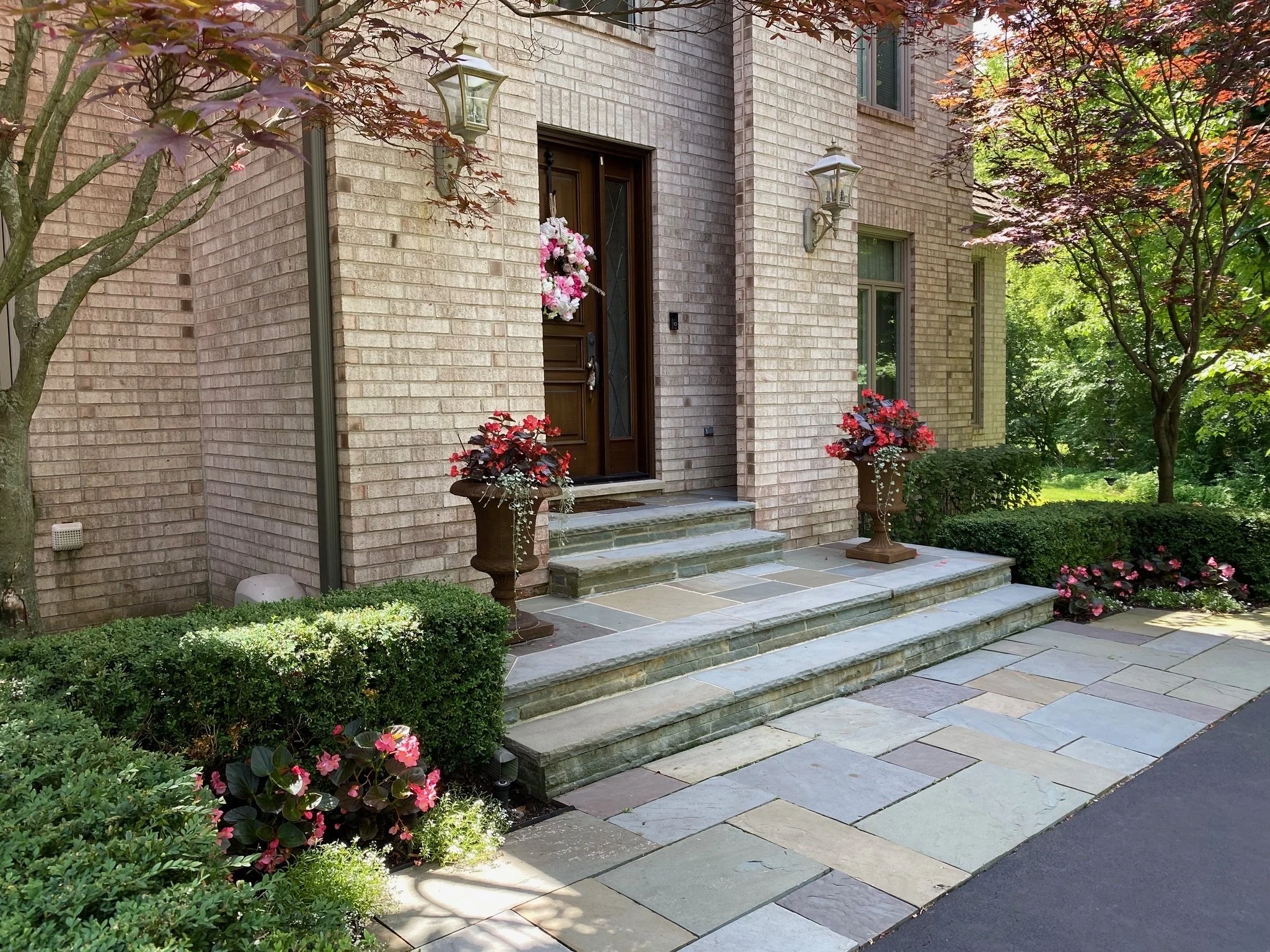 Front yard of a brick house with steps leading to the front door, decorated with flower pots and surrounded by greenery and trees.