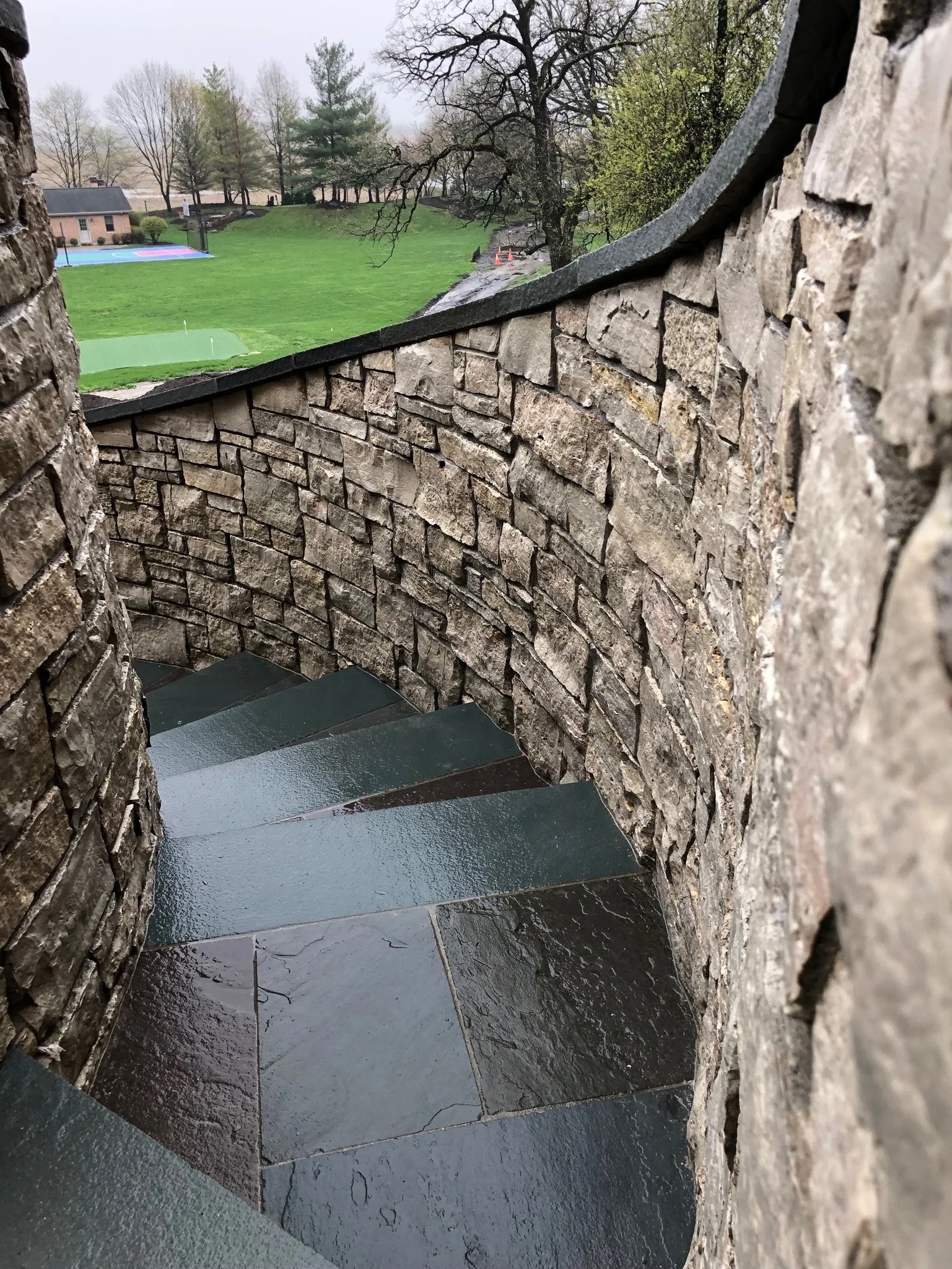 View through a circular stone opening of a curved staircase with dark steps, looking out onto a grassy yard with trees, a basketball court, and a house in the background.