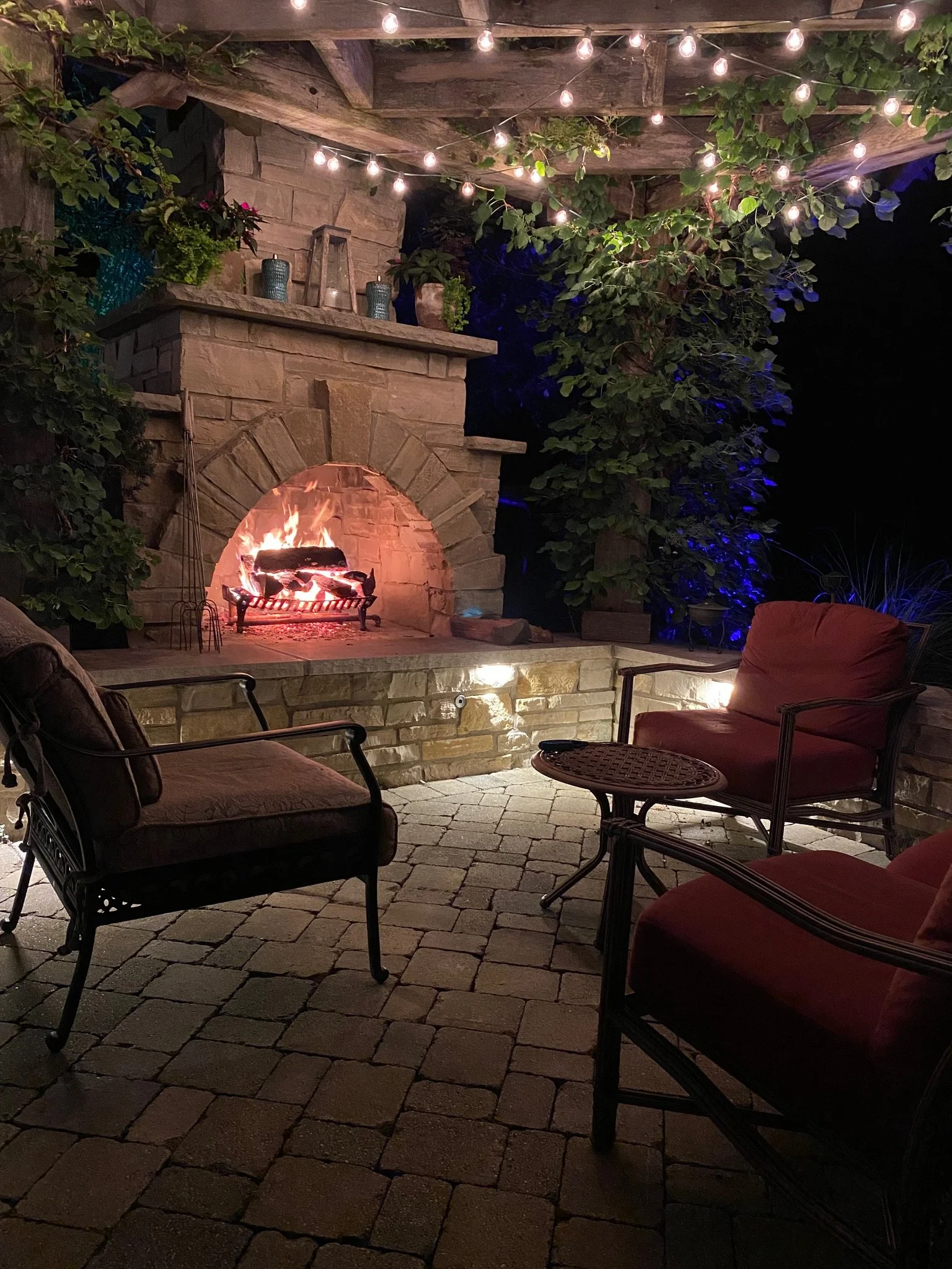 Outdoor patio area with a stone fireplace lit with a fire, surrounded by chairs with cushions, and decorated with string lights and greenery.